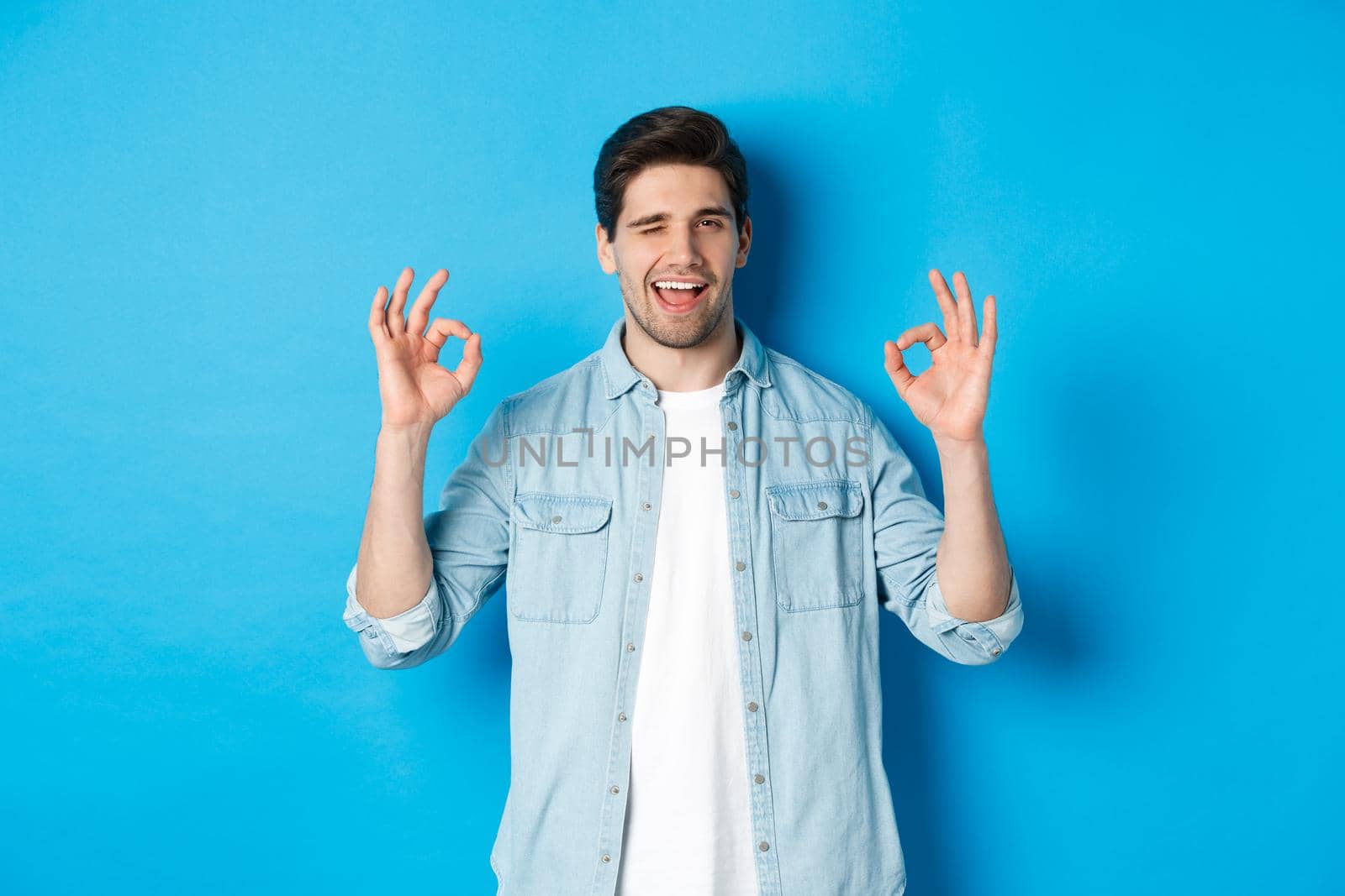 Relaxed and confident man showing ok signs and winking, everything okay gesture, standing against blue background.