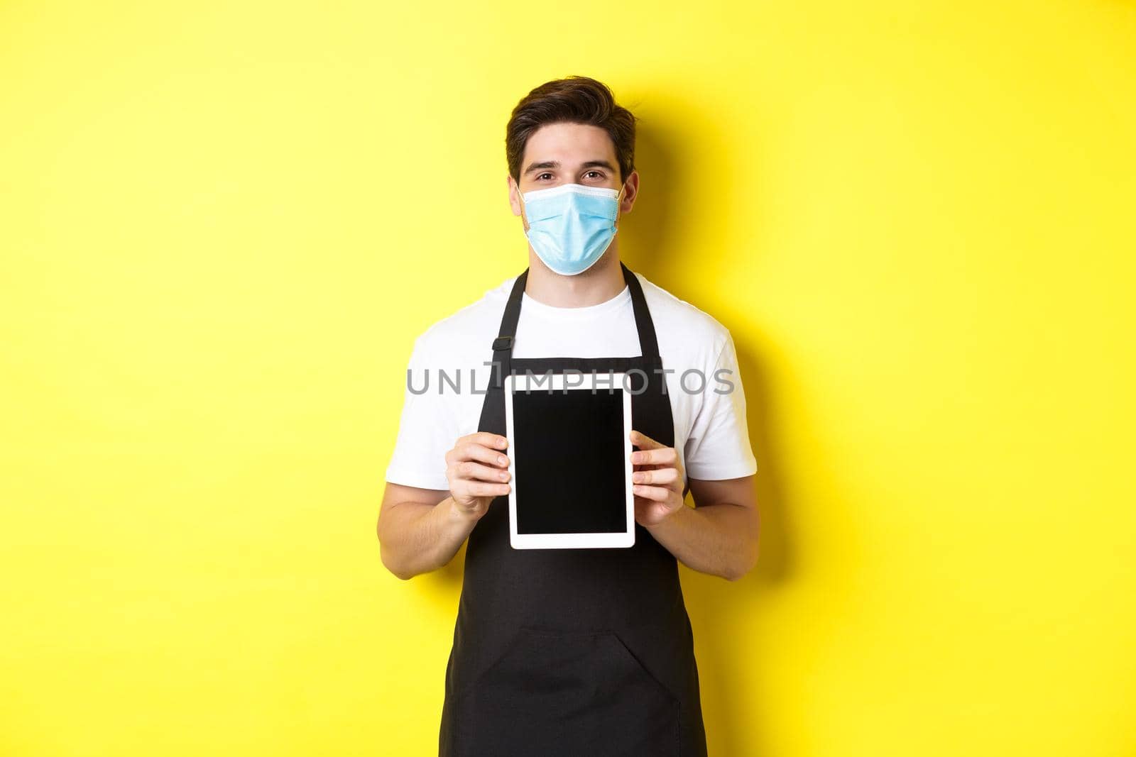 Concept of covid-19, small business and pandemic. Smiling barista in medical mask showing tablet screen, standing over yellow background by Benzoix