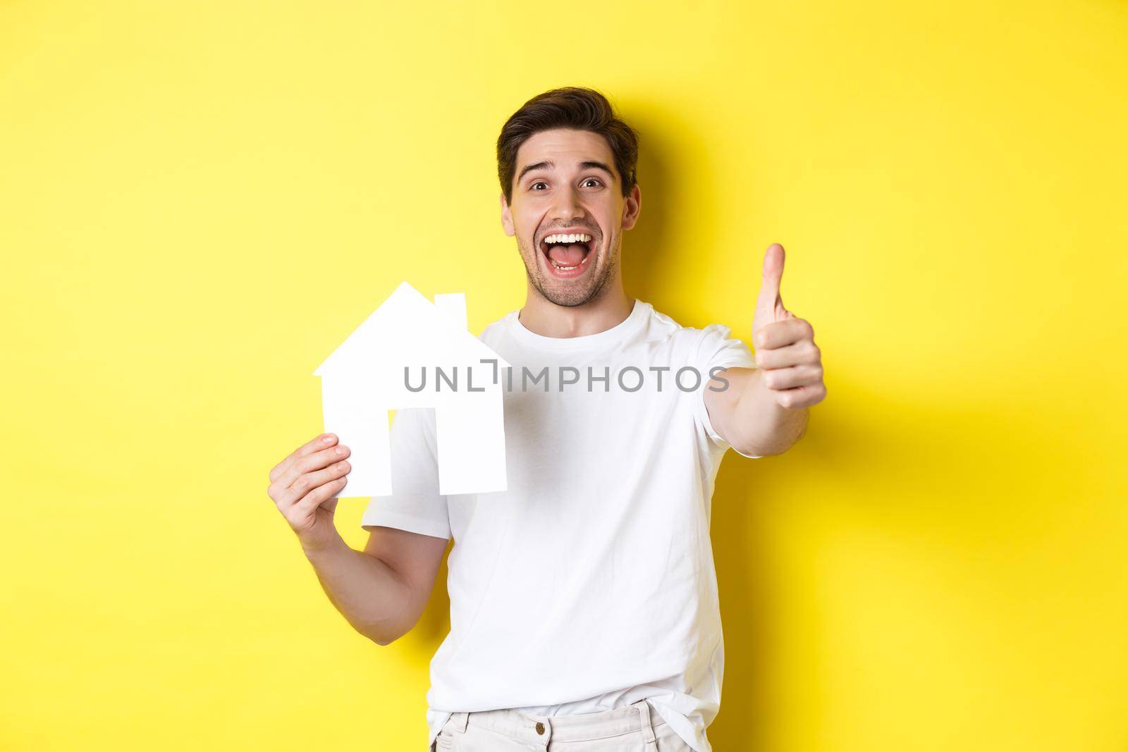 Real estate concept. Happy young male buyer showing thumb up and paper house model, smiling satisfied, standing over yellow background by Benzoix
