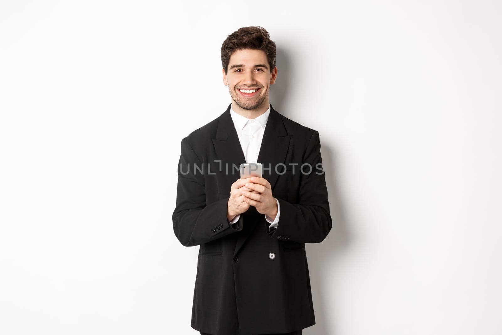 Portrait of successful, handsome businessman in suit, writing message on smartphone and smiling, standing over white background.