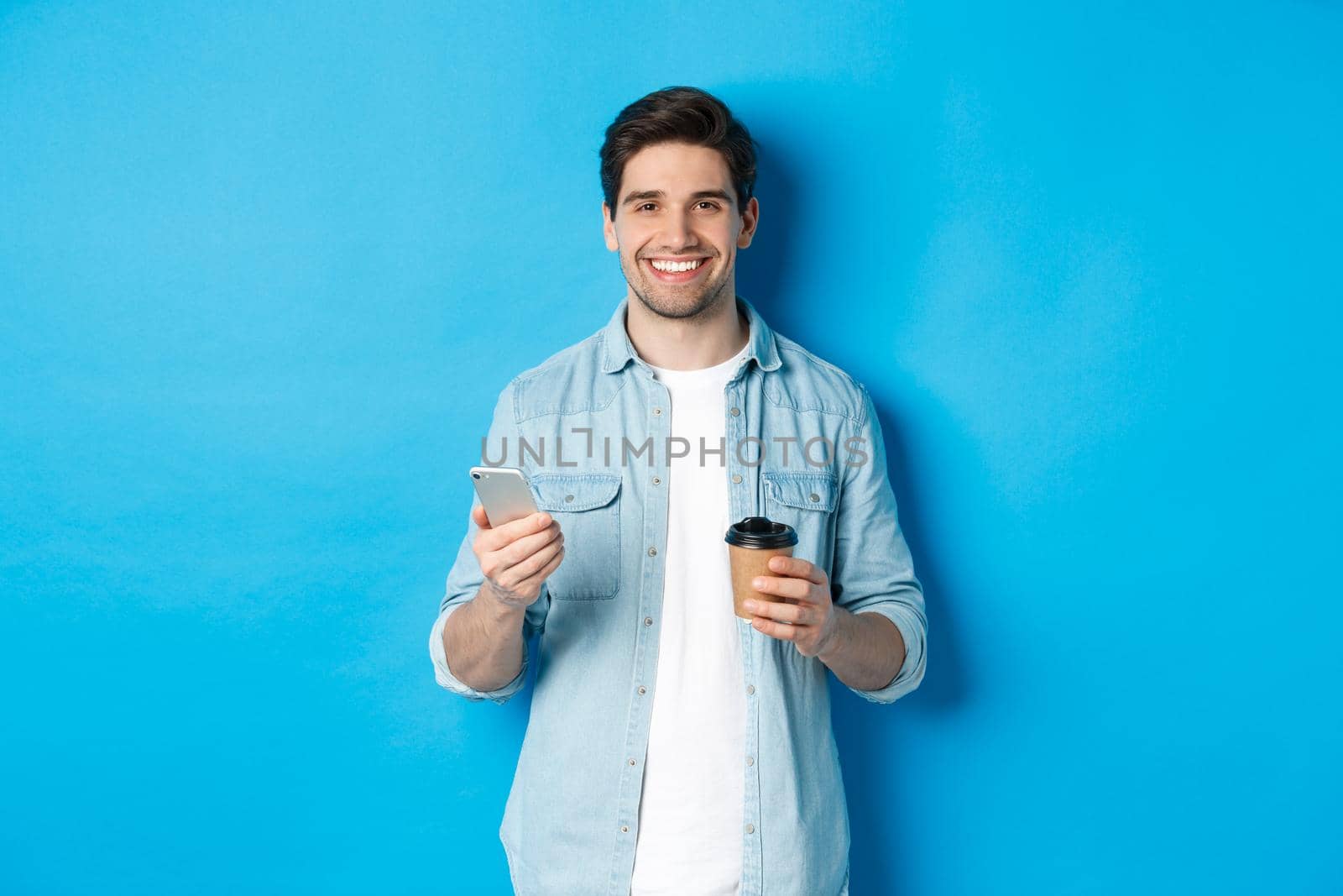 Young modern man drinking coffee and using mobile phone, smiling at camera, standing over blue background.