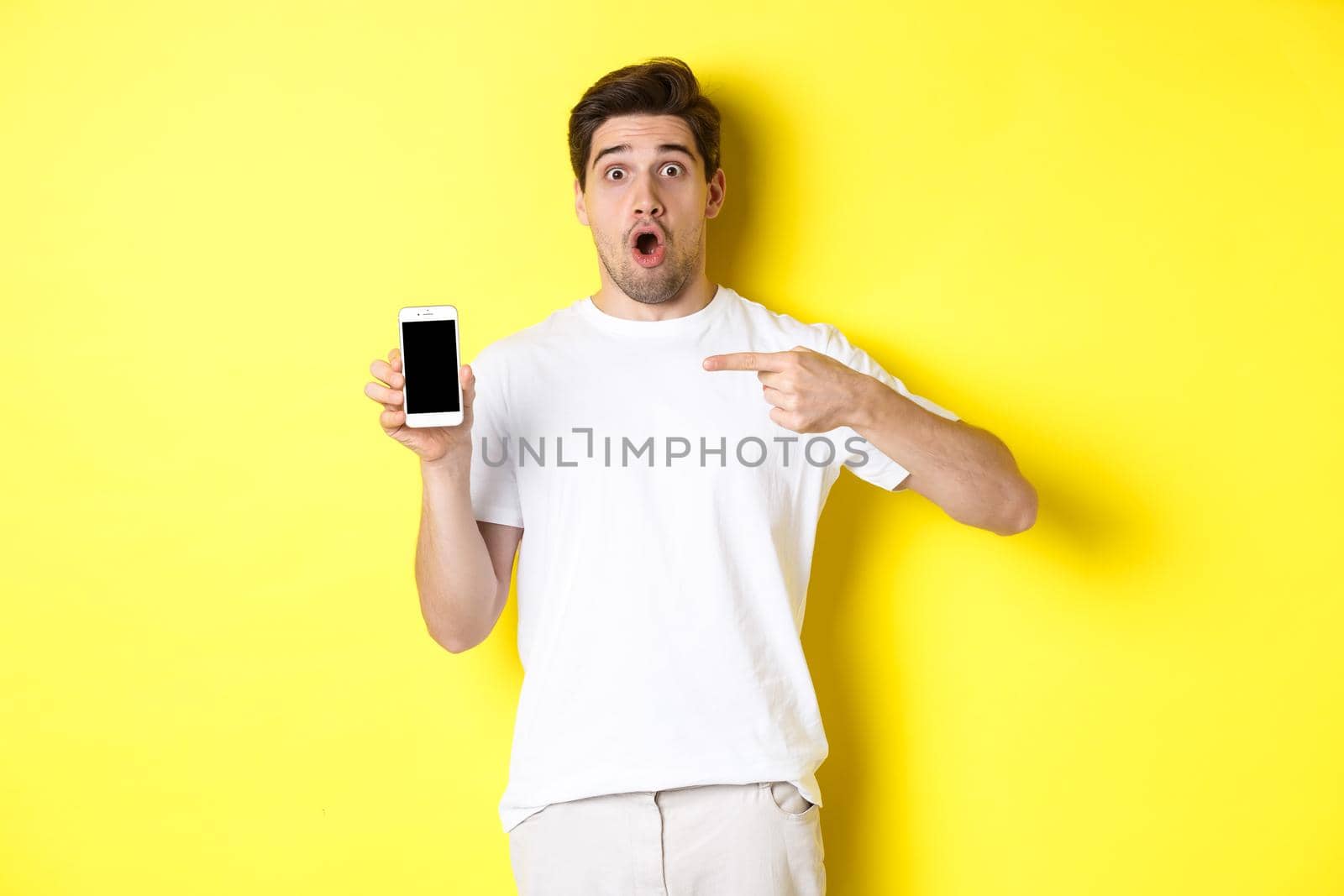 Impressed guy check out smartphone screen, pointing finger at display and gasping amazed, standing over yellow background by Benzoix