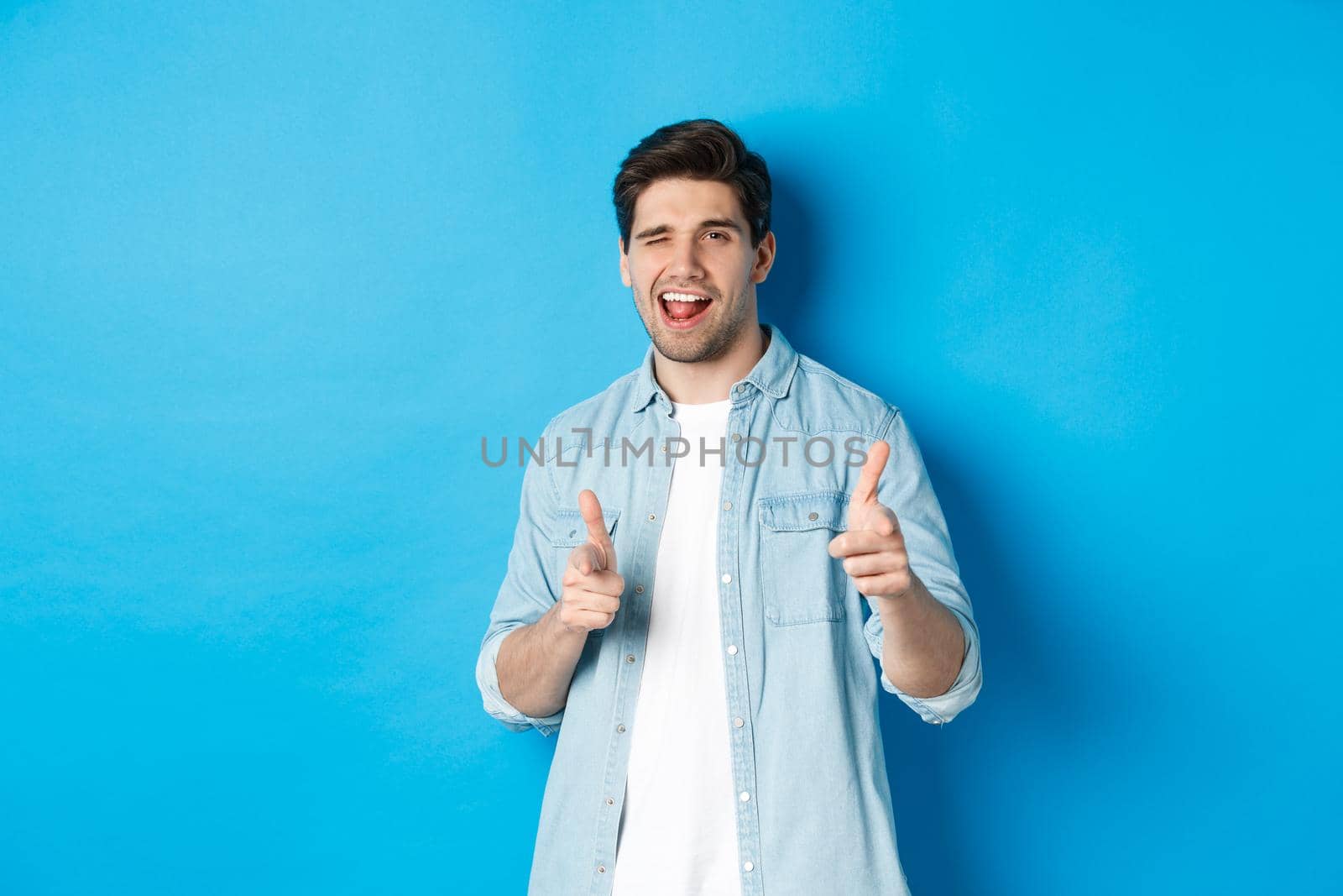 Confident adult man winking, pointing finger at you and winking, congratulating or praising person, standing against blue background.