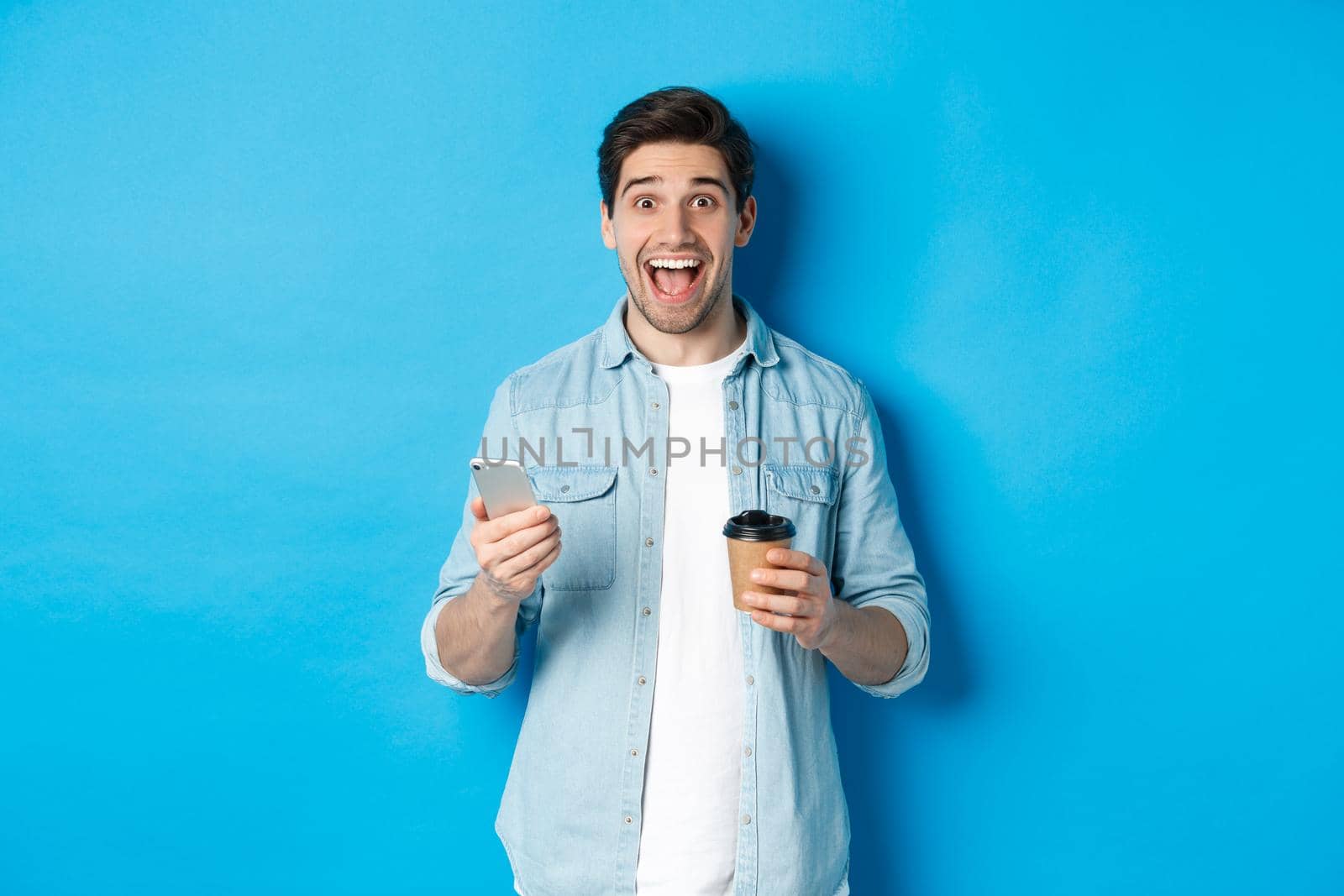 Happy young man drinking coffee and using mobile phone, looking excited, standing against blue background.