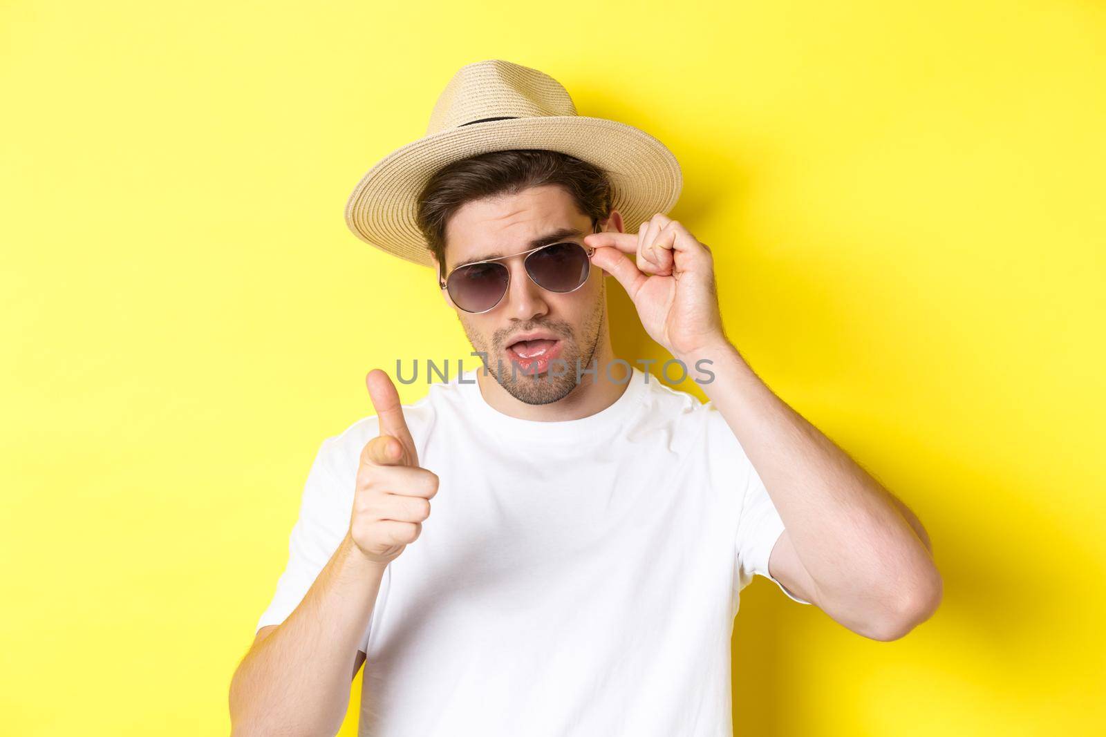 Concept of tourism and vacation. Cool and sassy man flirting with you, wearing sunglasses and pointing finger at camera, standing over yellow background by Benzoix