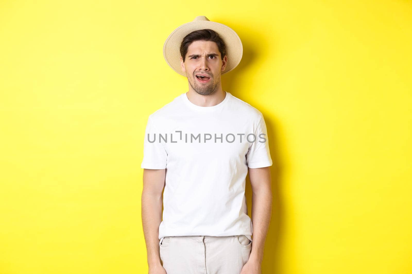 Concept of tourism and summer. Confused guy traveller in straw hat, looking puzzled, cant understand something, standing over yellow background.