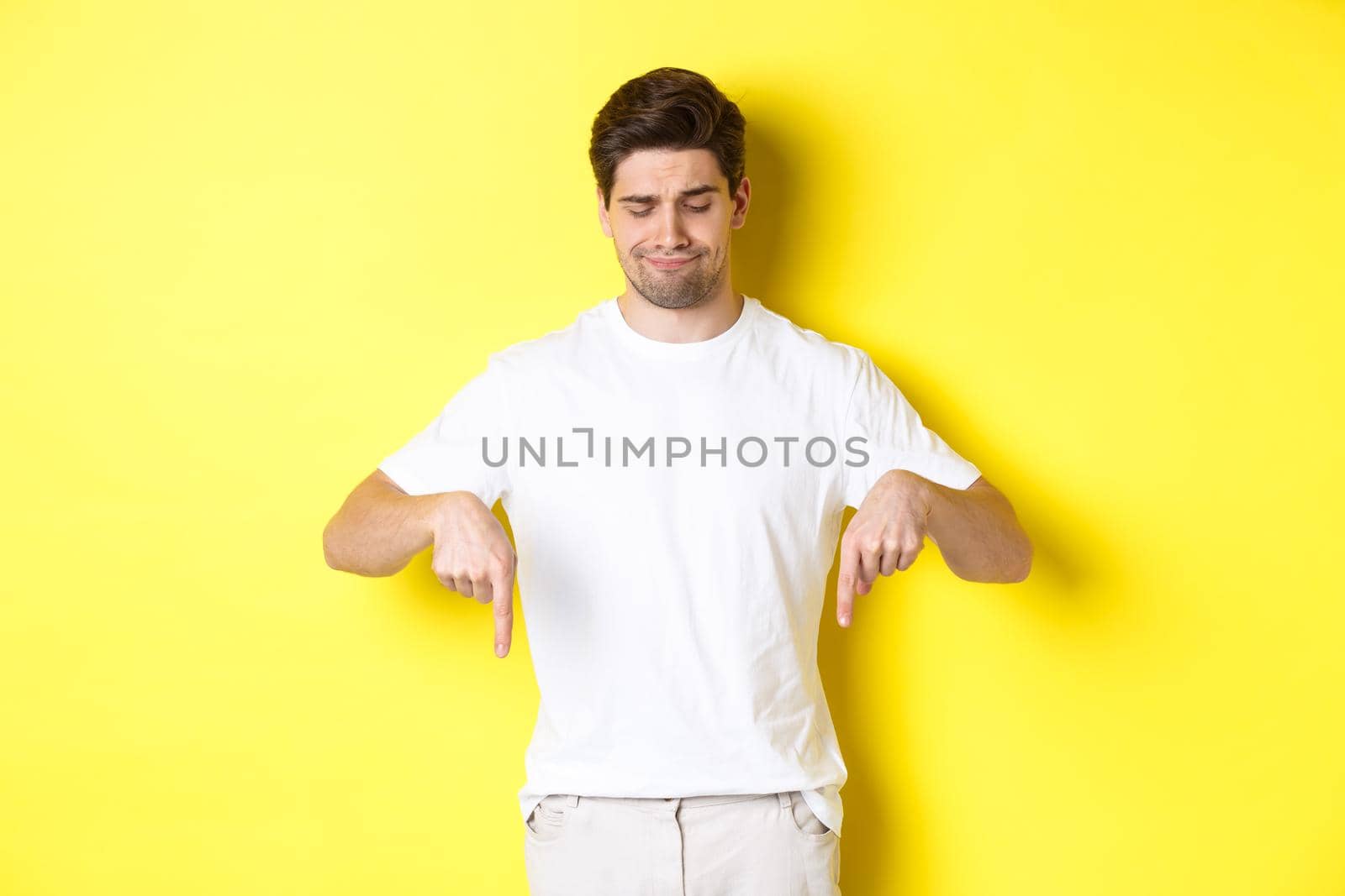 Skeptical young man in white t-shirt, pointing and looking down upset, disapprove and dislike product, standing over yellow background by Benzoix