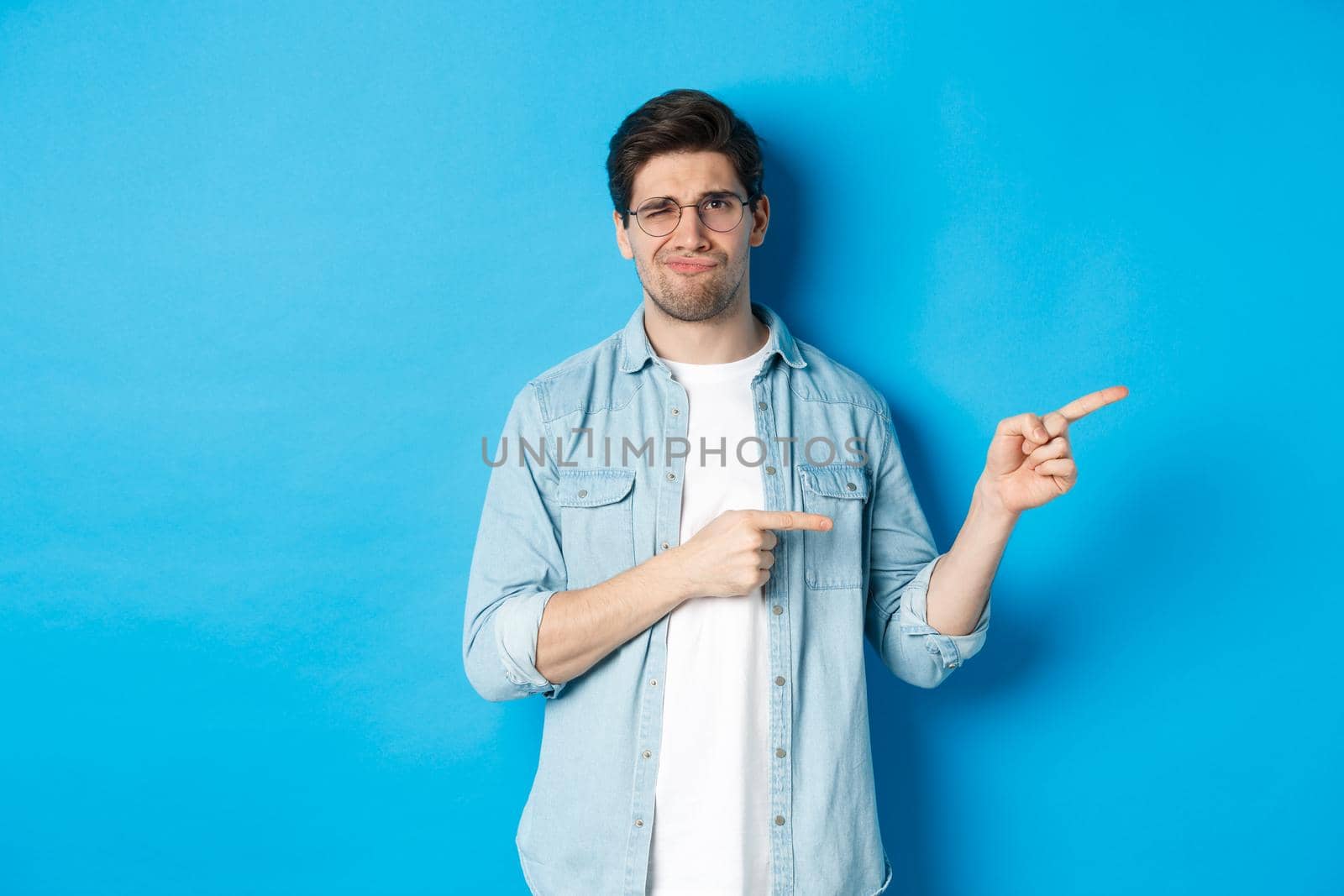 Disappointed young man in glasses pointing fingers right at copy space, showing promo banner and smirking dissatisfied, judging bad product, standing over blue background.