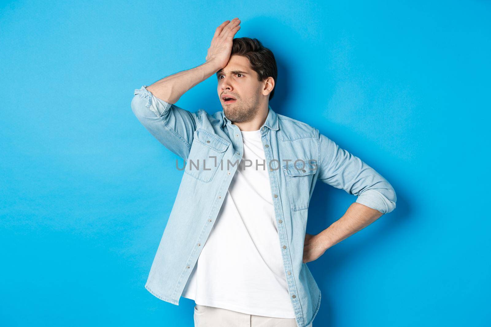 Upset man making facepalm and looking away concerned, forgot something important, slap forehead troubled, standing against blue background by Benzoix