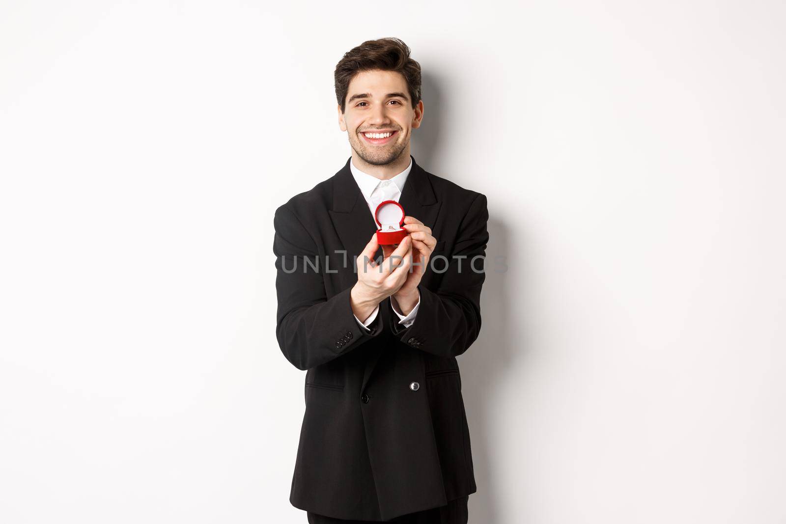 Image of handsome boyfriend in black suit making a proposal, asking to marry him and showing wedding ring, standing over white background.