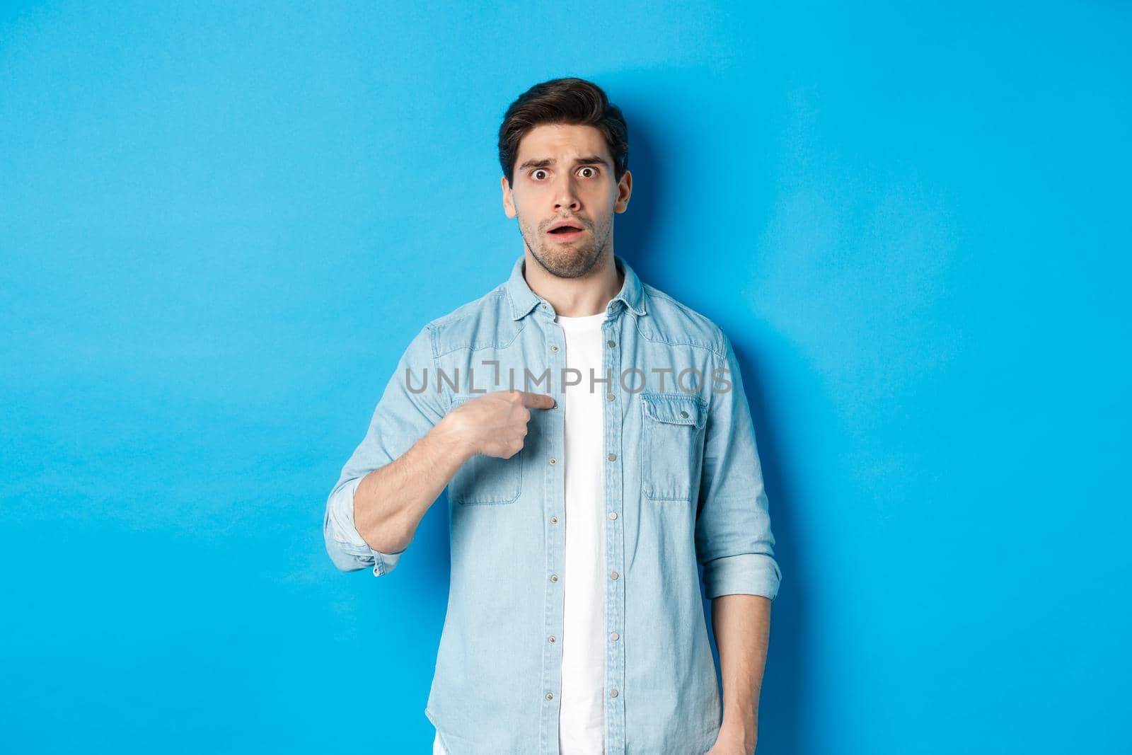 Nervous man pointing at himself and looking confused, standing in casual clothes over blue background.