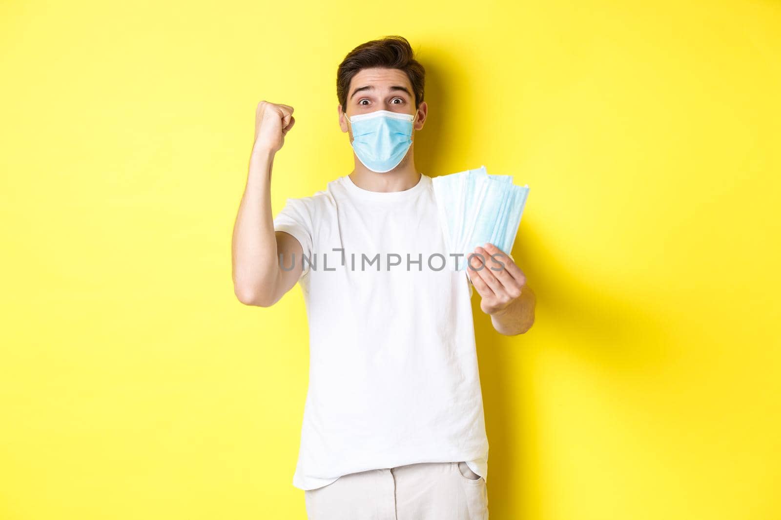 Concept of covid-19, quarantine and preventive measures. Happy man triumphing, raising hand to celebrate something and giving medical mask, standing against yellow background by Benzoix