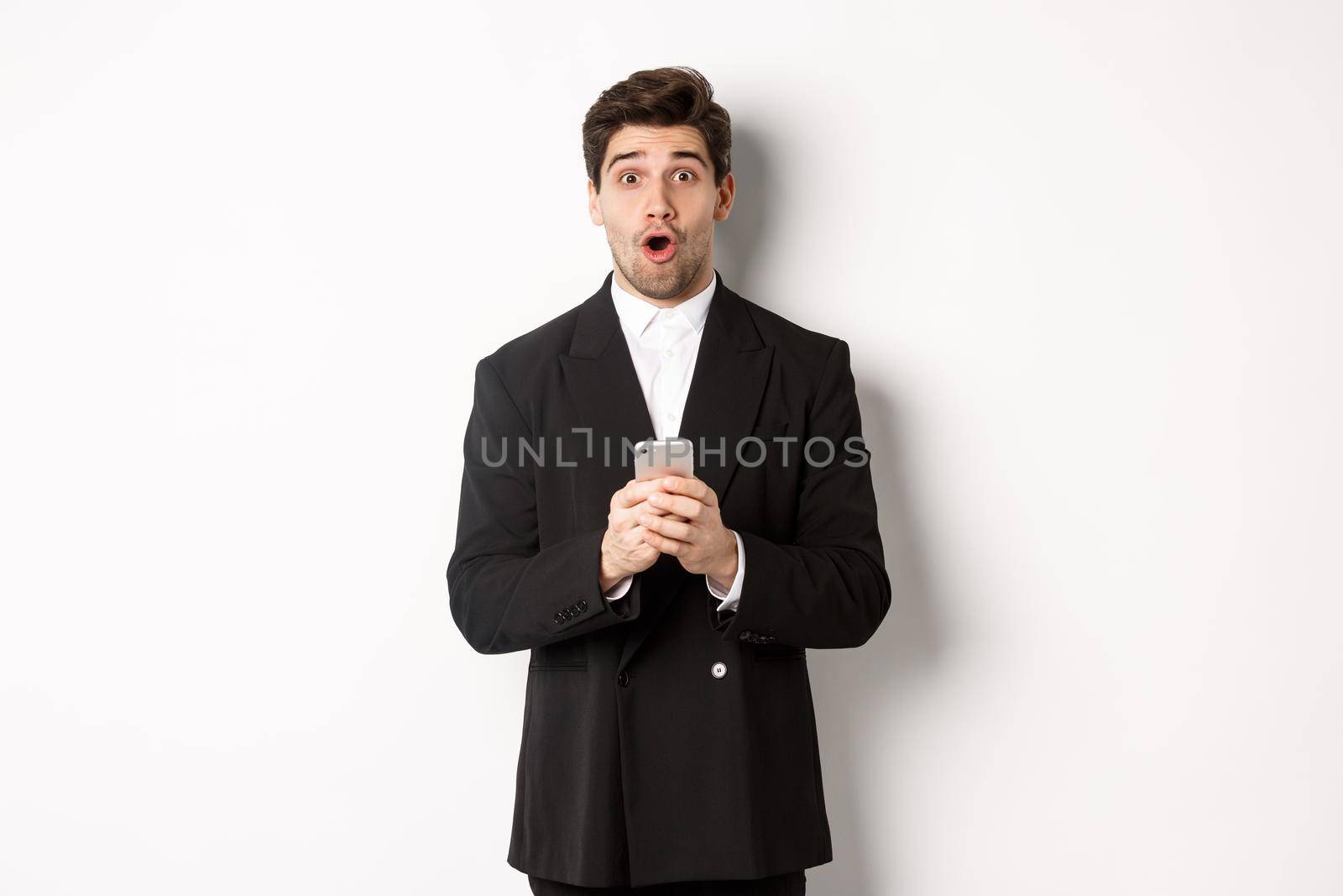 Amazed handsome guy in black suit reacting to cool promo offer, holding mobile phone, standing against white background.