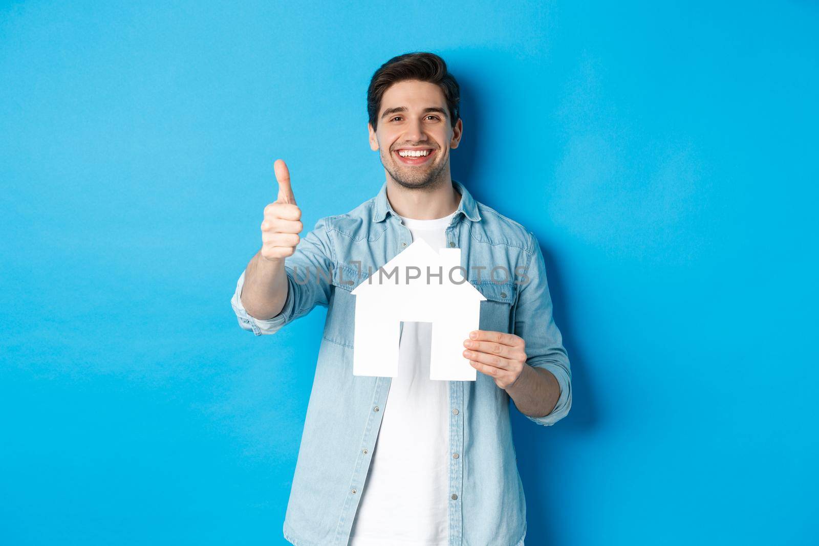 Insurance, mortgage and real estate concept. Satisfied client showing house model and thumb up, smiling pleased, standing against blue background.