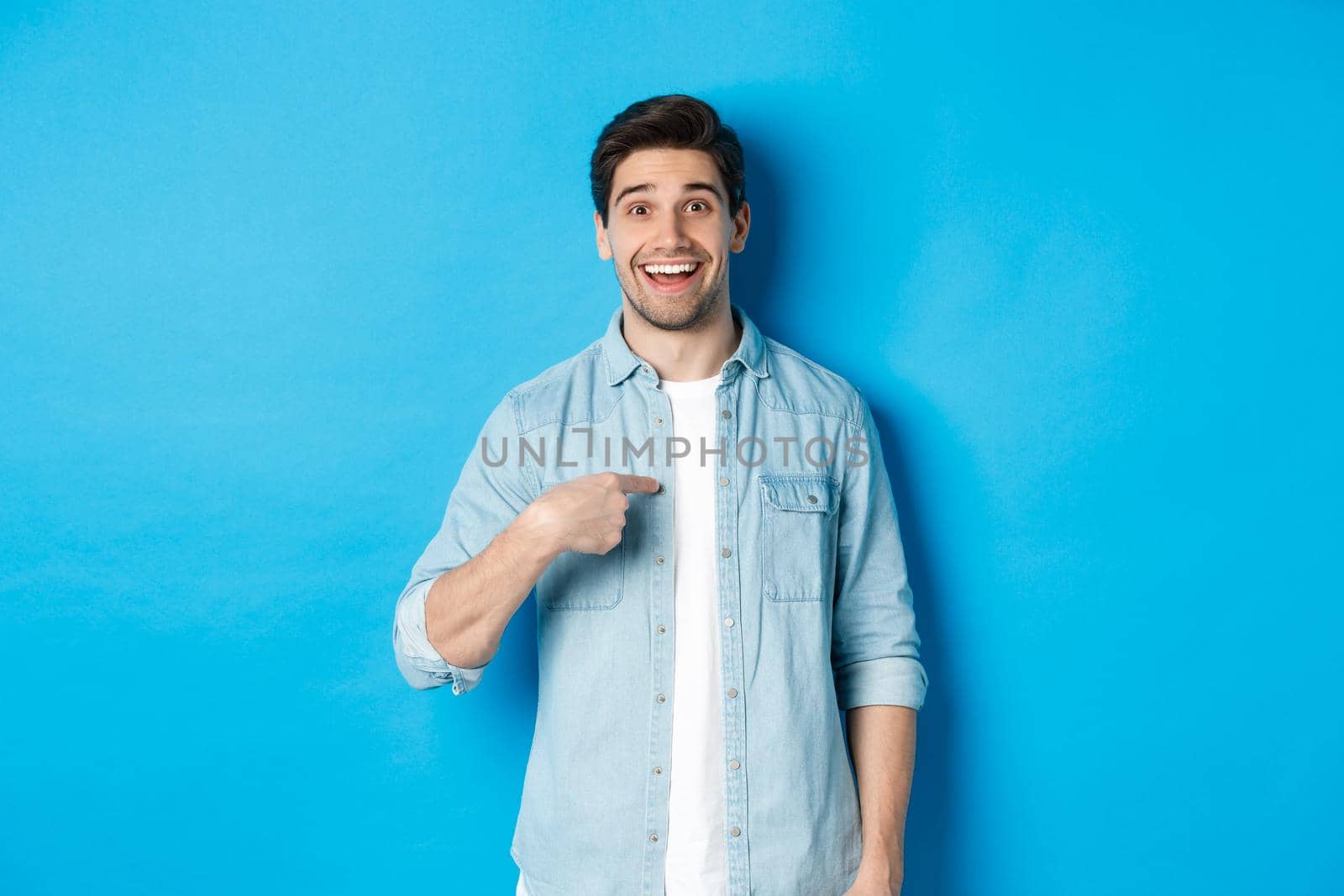 Happy and surprised man pointing at himself, smiling pleased, standing against blue background.