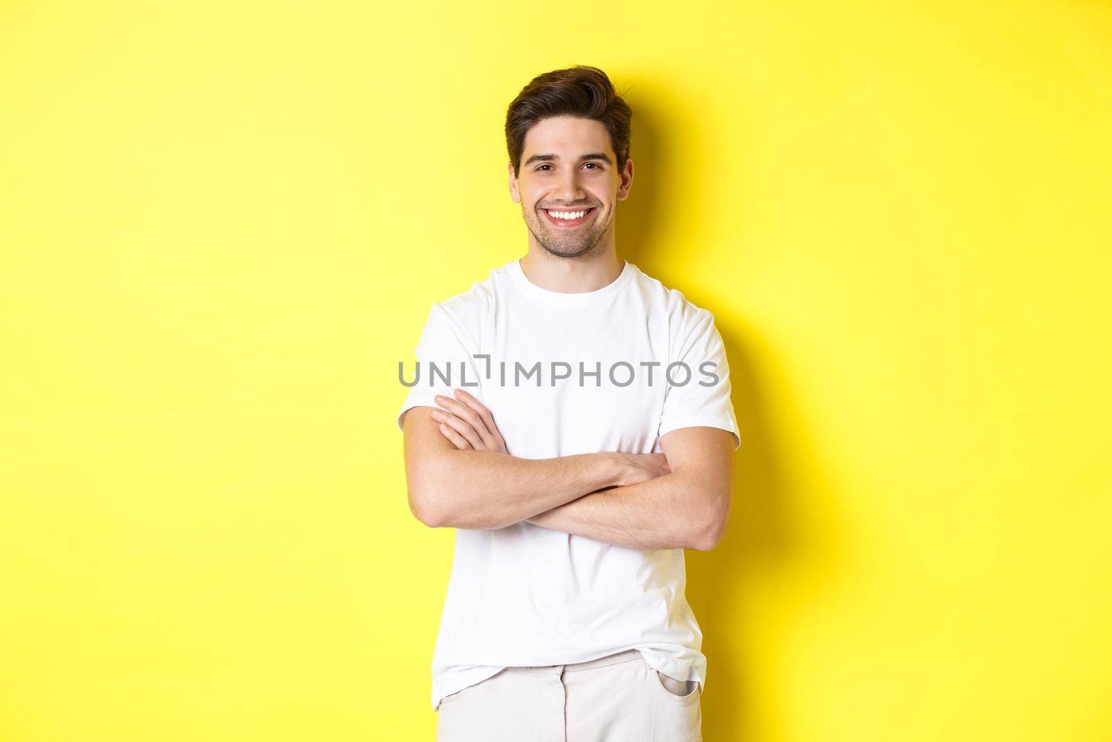 Image of confident caucasian man smiling pleased, holding hands crossed on chest and looking satisfied, standing over yellow background.