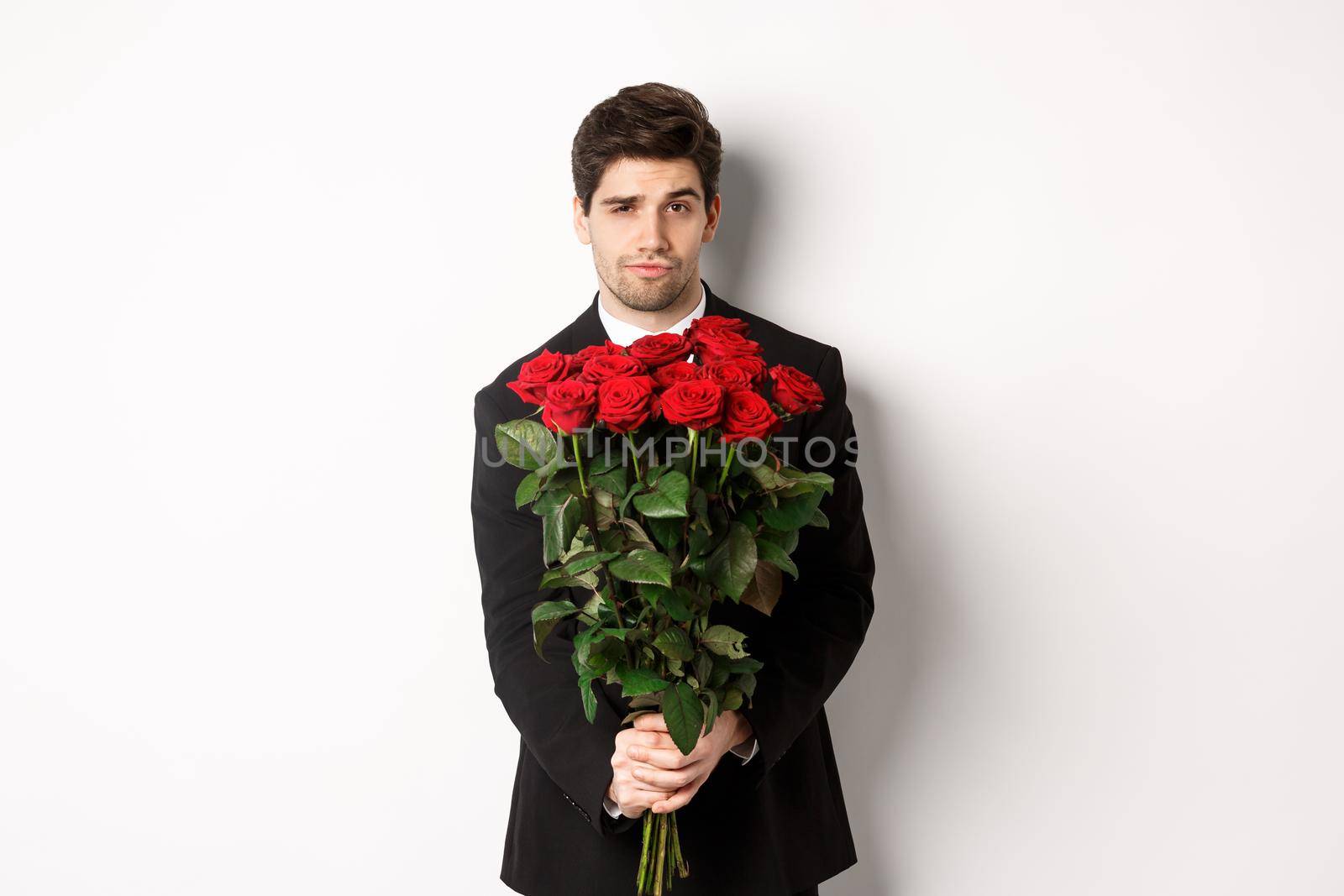 Image of handsome boyfriend in black suit, holding bouquet of red roses and smiling, being on a date, standing over white background.