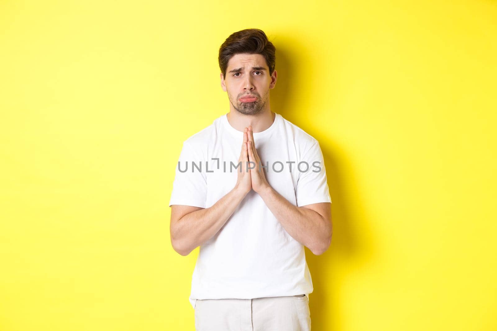 Sad guy begging for something, sulking and asking for favour, saying sorry, standing over yellow background.