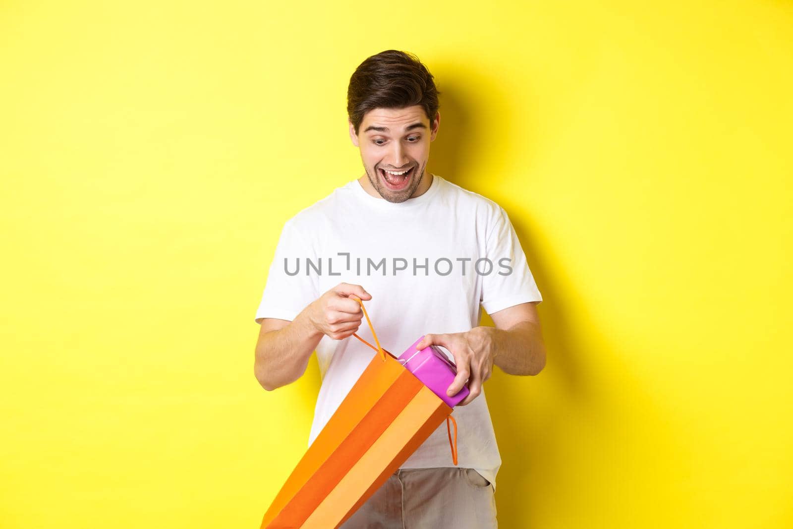 Concept of holidays and celebration. Young man looking surprised as take out gift from shopping bag, standing over yellow background by Benzoix