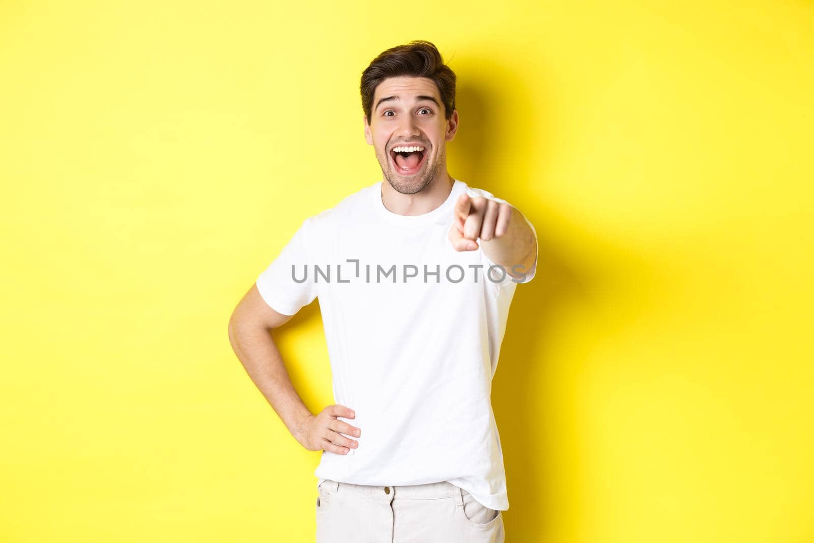 Happy guy pointing finger at camera and laughing, check out something, standing over yellow background.
