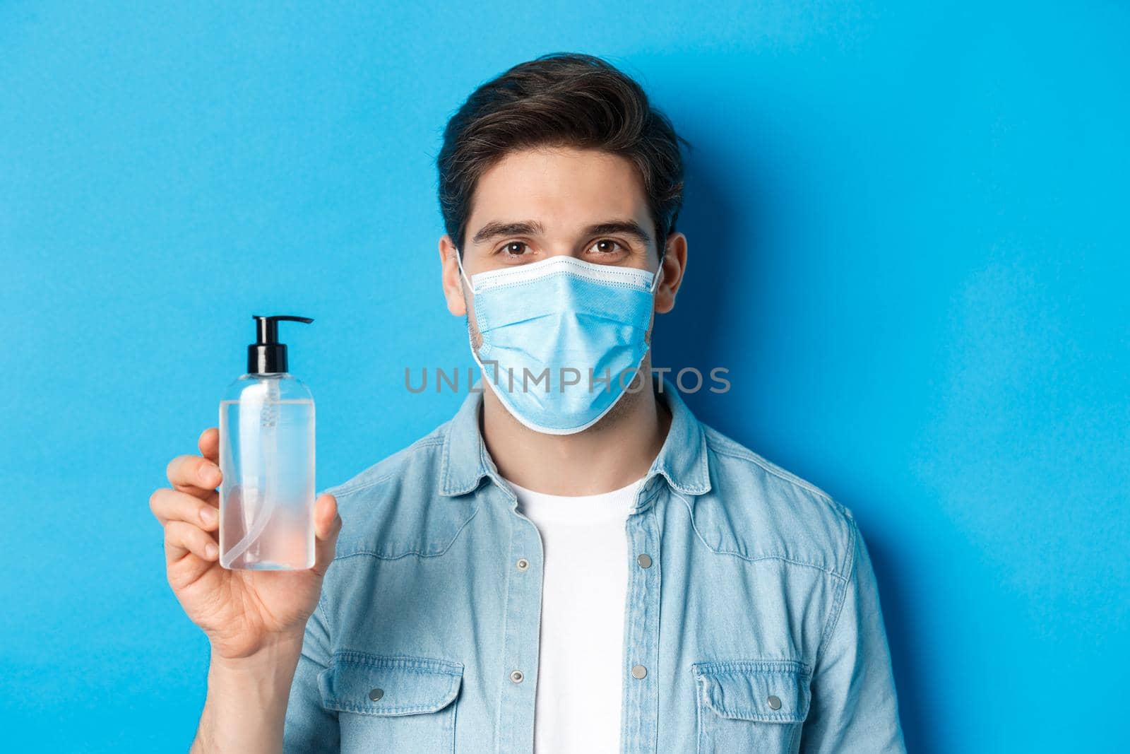 Concept of coronavirus, pandemic and preventive measures. Close-up of young man in medical mask advice to use hand sanitizer, showing antiseptic bottle, standing over blue background.