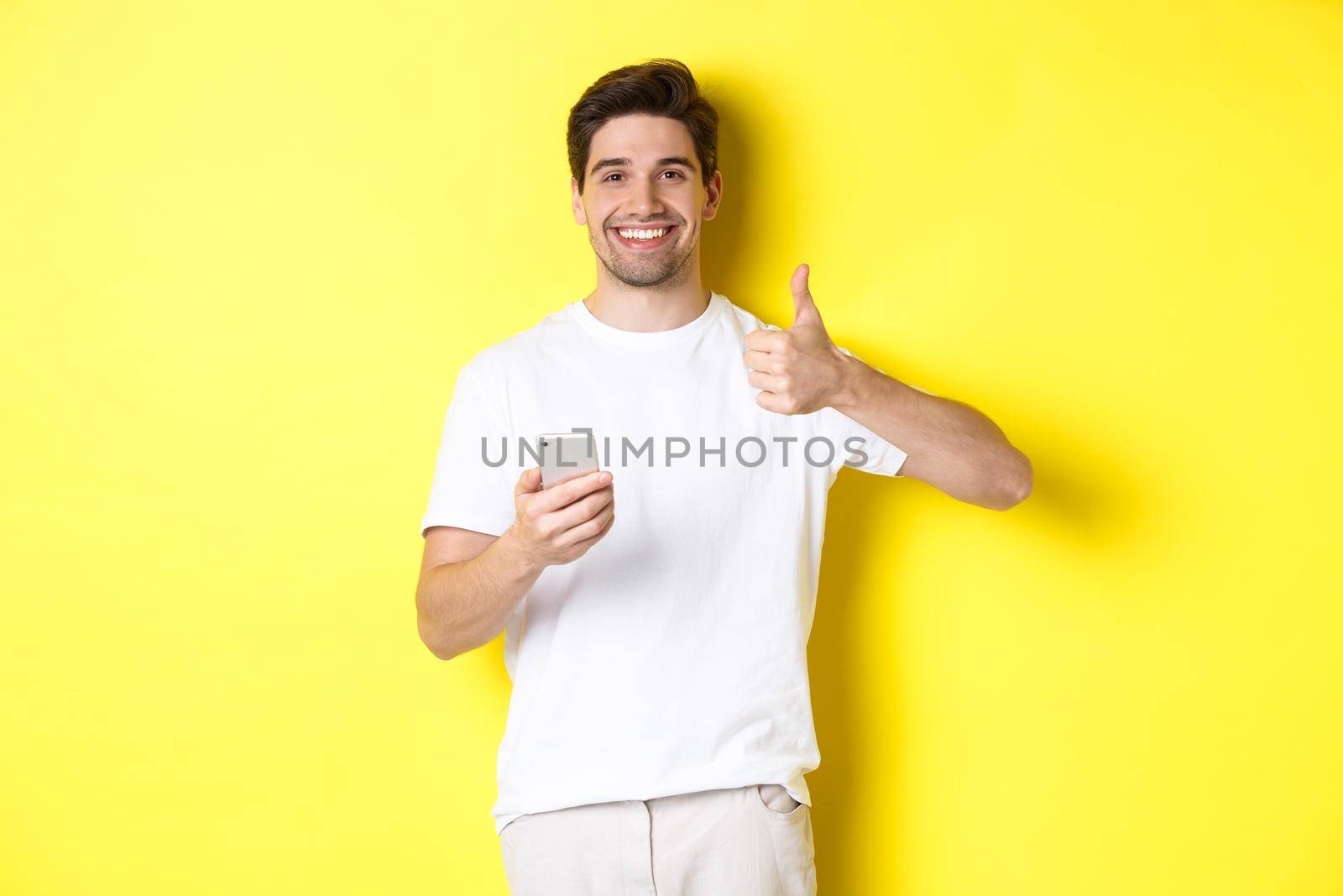 Happy satisfied man holding smartphone, showing thumb up in approval, recommend something online, standing over yellow background.