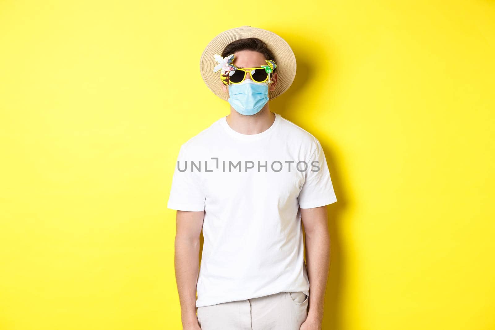 Concept of covid-19, vacation and social distancing. Man tourist wearing medical mask and summer hat with sunglasses, going on trip during pandemic, yellow background.