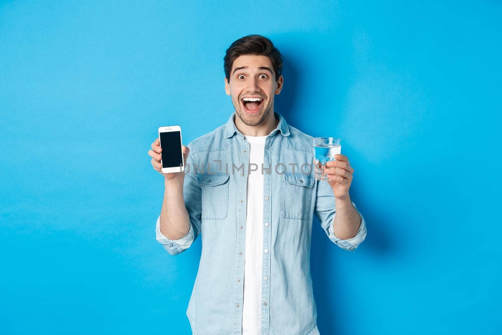 Happy man showing glass of water and mobile screen, recommending smartphone health app, standing over blue background.