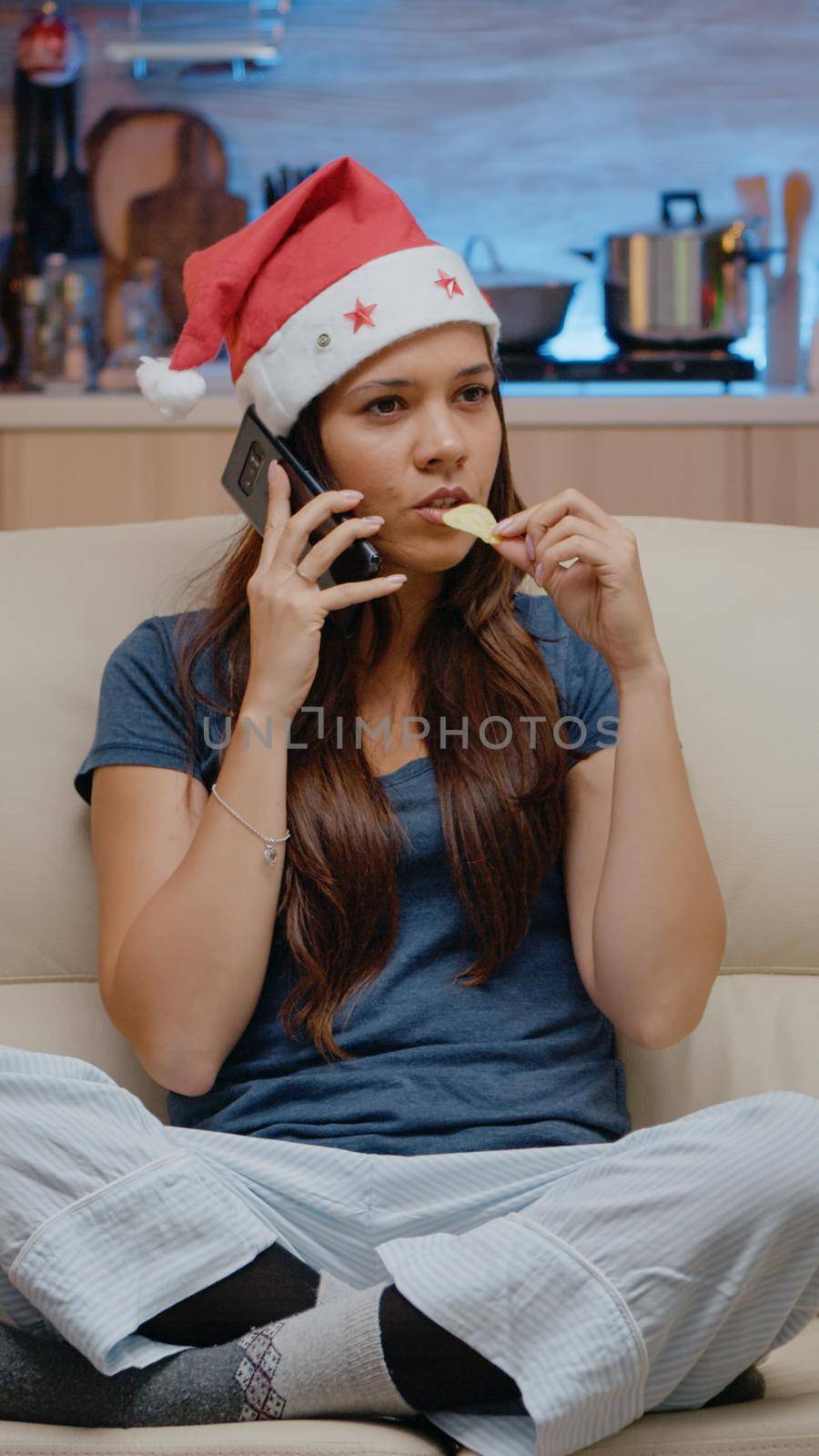 Festive woman talking on smartphone with family for remote christmas celebration. Adult with red santa hat using call on phone chatting while watching television and eating chips