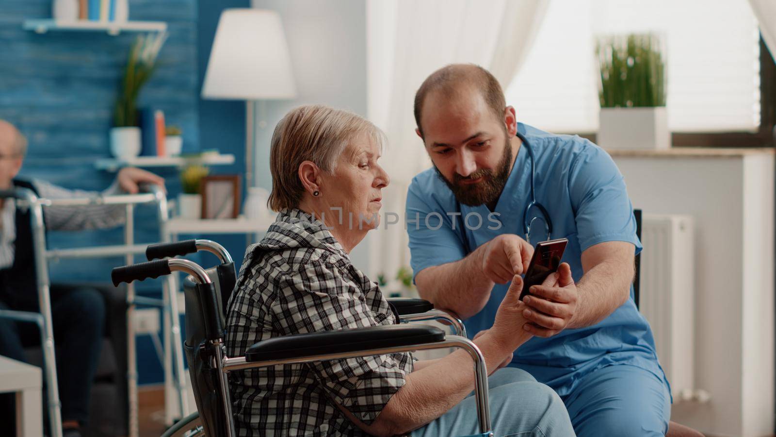 Man nurse teaching retired woman with disability to use smartphone for communication in nursing home. Senior patient sitting in wheelchair learning phone technology with assistance