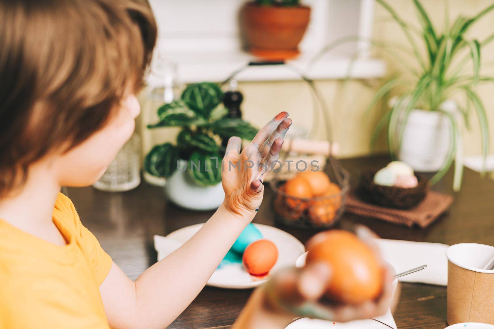 Easter day. Cropped close up Kid boy with dirty hands painting eggs on wooden background. Child sitting in a kitchen. Preparing for Easter, creative homemade decoration.