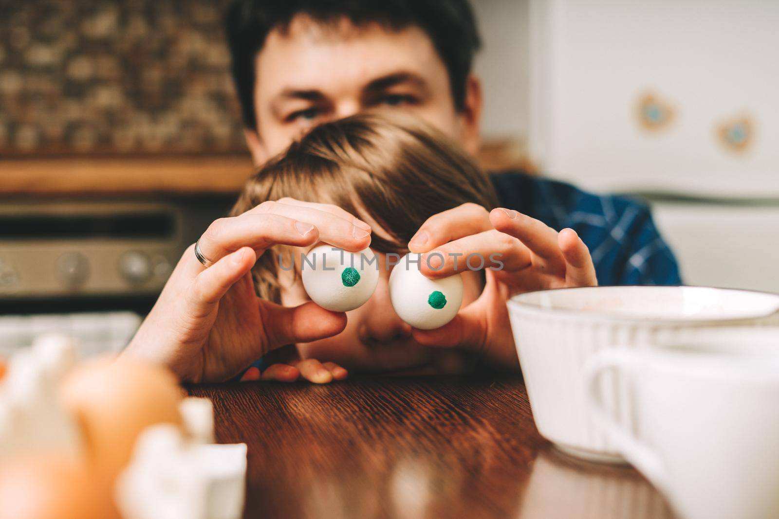 Easter day with kid. Father and son with funny face and eggs eyes, having fun together. Family man and child in a kitchen. Preparing for Easter, creative homemade decoration.