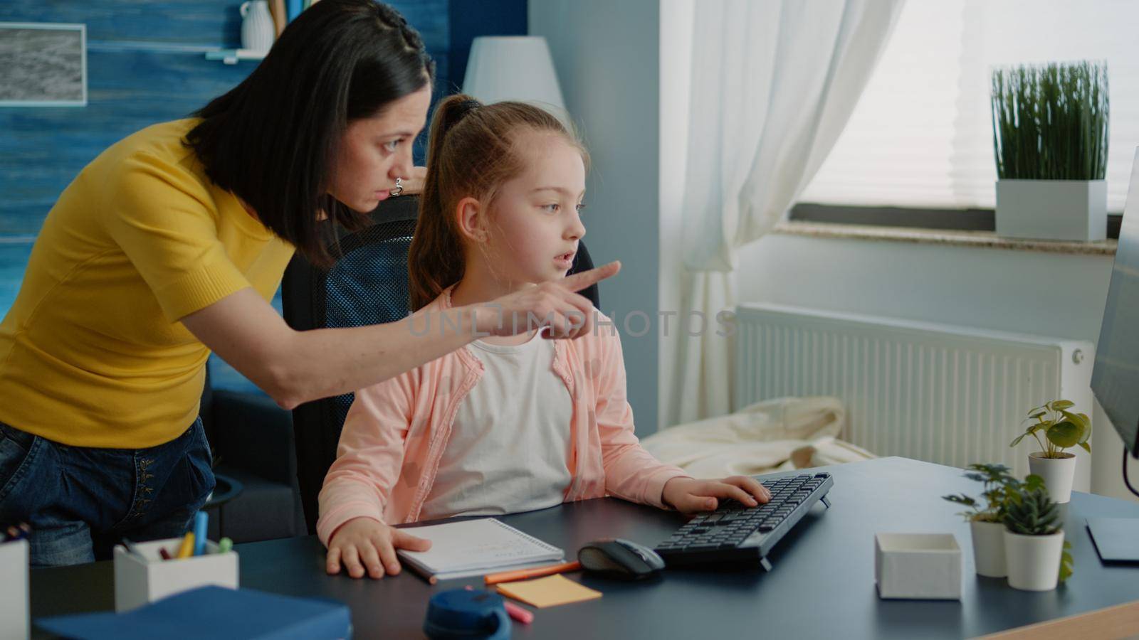 Mother helping young child with homework for remote education and knowledge. Schoolgirl receiving assistance from parent while using computer and notebook for online class lesson.