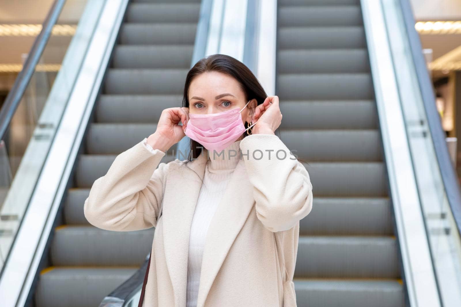 beautiful woman putting on pink medical mask on her face against the background of the escadator in a public place