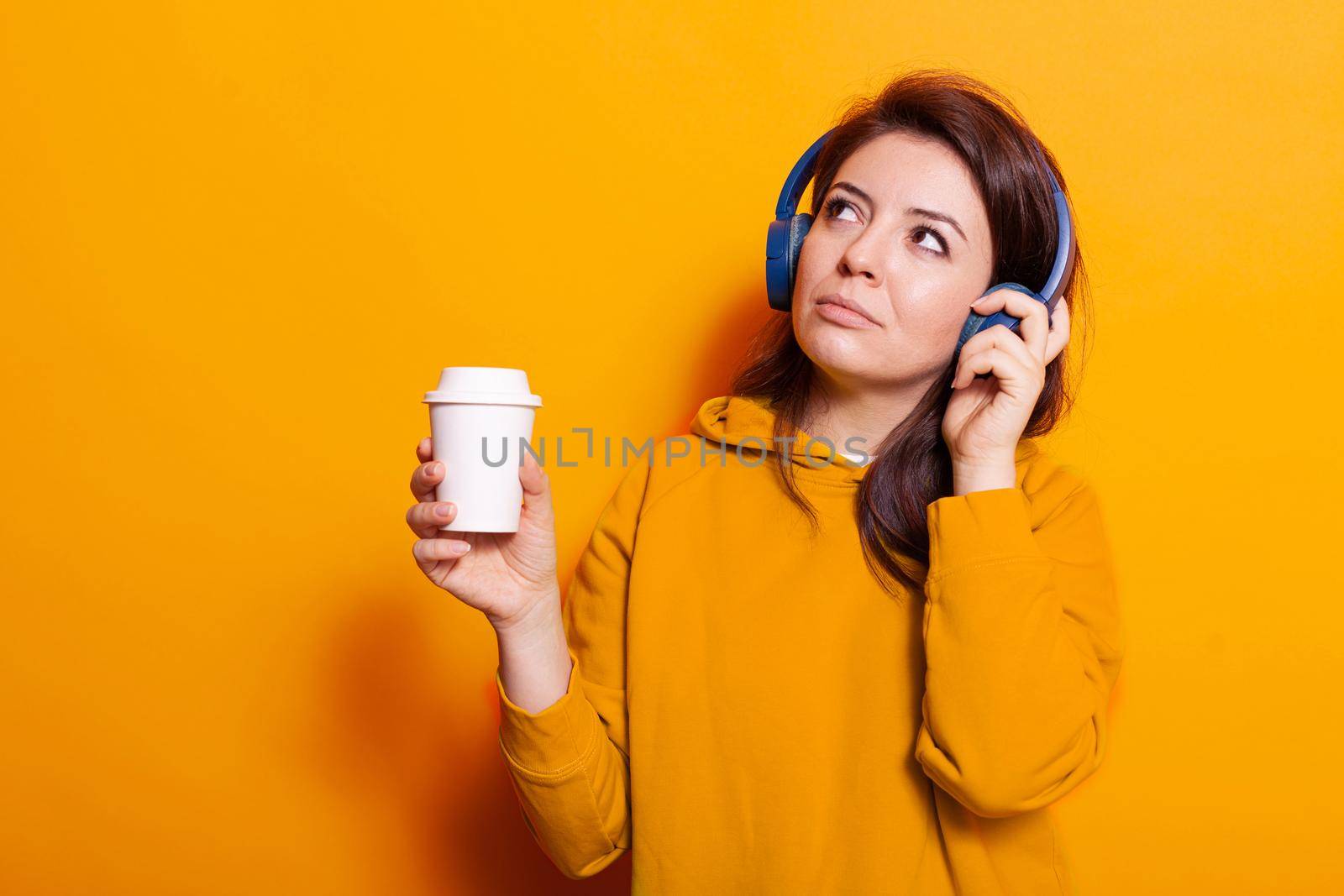 Happy person with cup of coffee listening to music on headset. Caucasian woman feeling relaxed and enjoying song on headphones while holding hot beverage to drink. Young adult relaxing