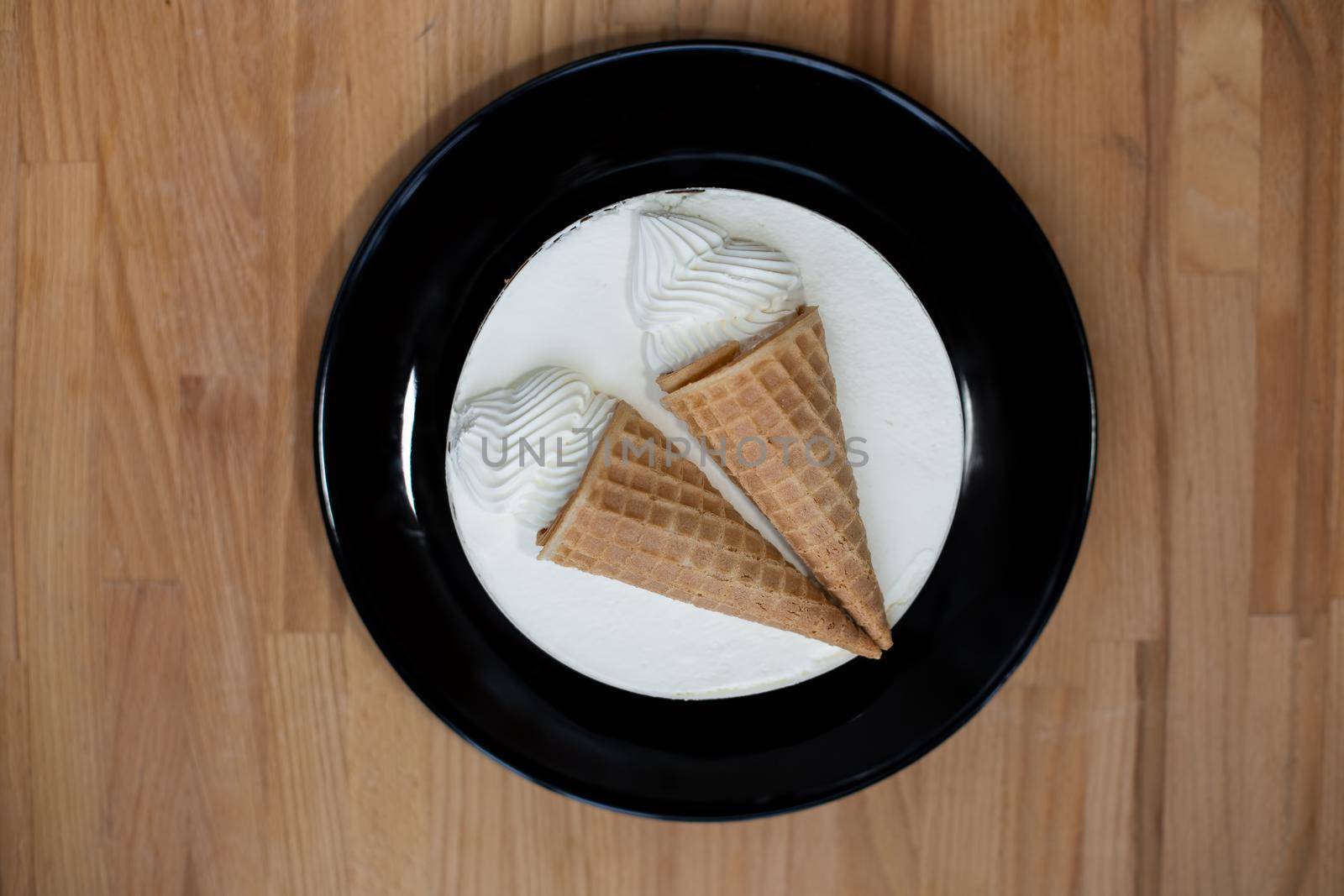 round white dessert sweet tasty ice cream cake with decoration two ice cream cones in a waffle cup on a black plate on a wooden board background. top view, close-up by Mariaprovector