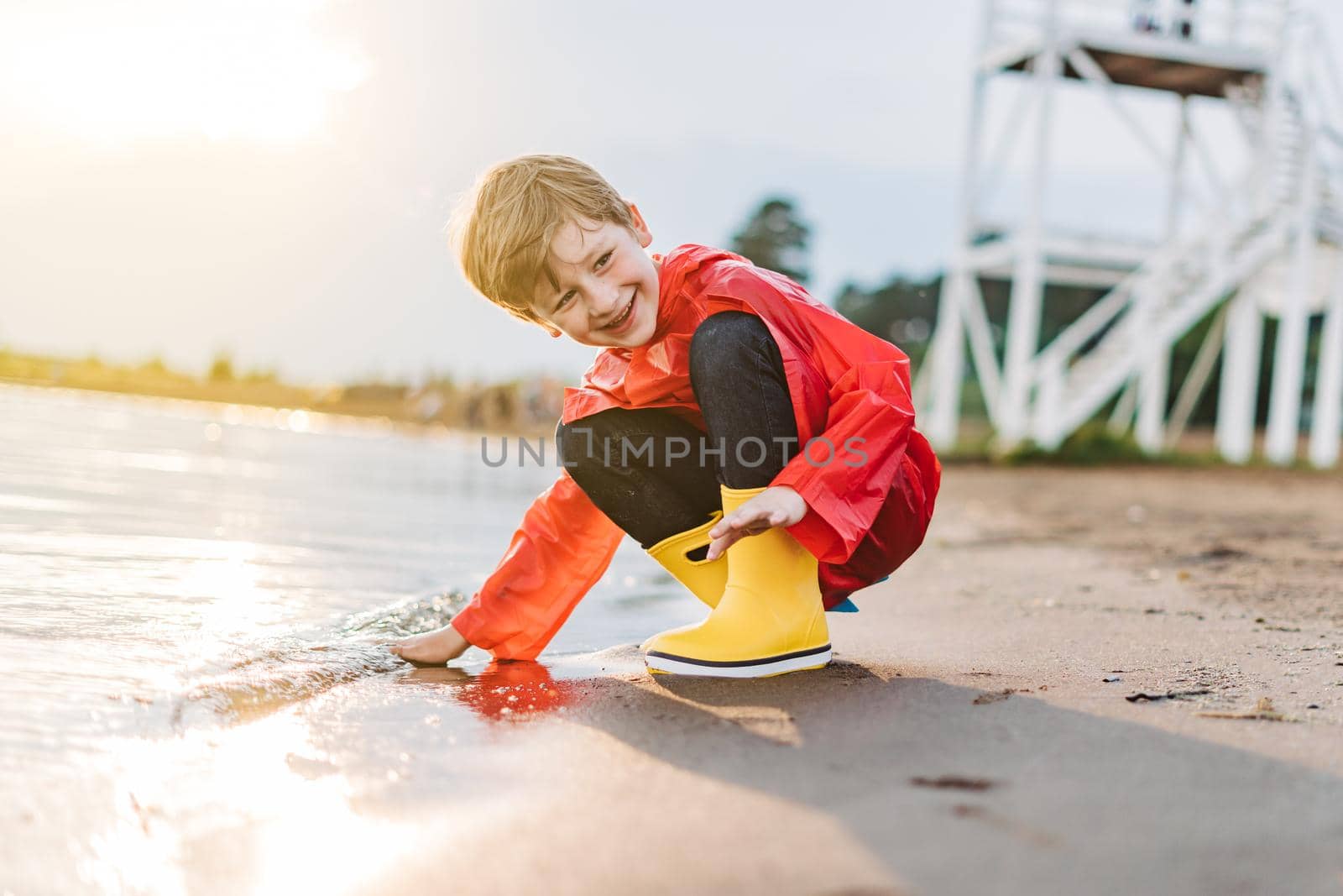Boy in a red raincoat and yellow rubber boots playing with water at the beach. School kid in a waterproof coat touching water at sea. Child having fun with waves at the shore.