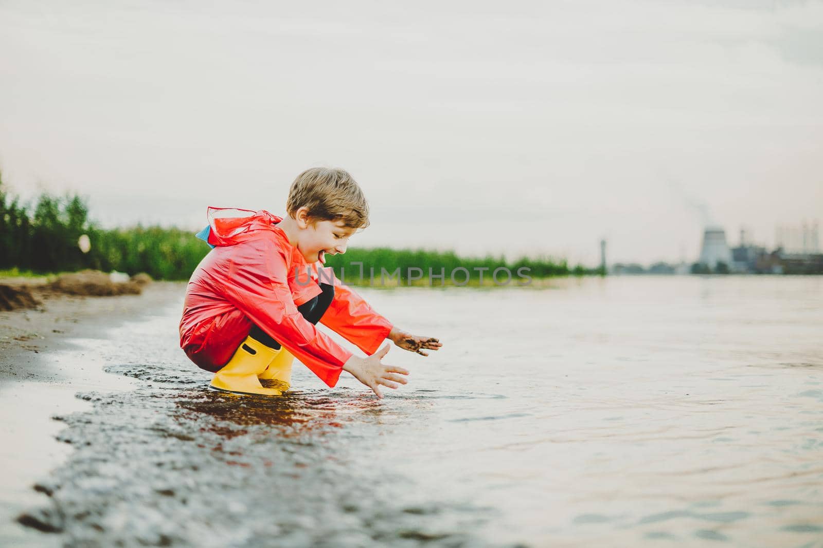 Boy in a red raincoat and yellow rubber boots playing with water at the beach. School kid in a waterproof coat touching water at sea. Child having fun with waves at the shore.