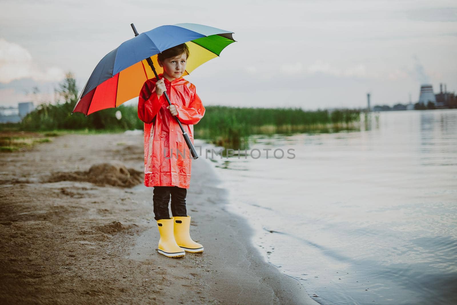 Boy in a red raincoat and yellow rubber boots stands at river bank and holding rainbow umbrella. School kid standing still near autumn lake. Child wearing waterproof clothes at shoreside by Ostanina