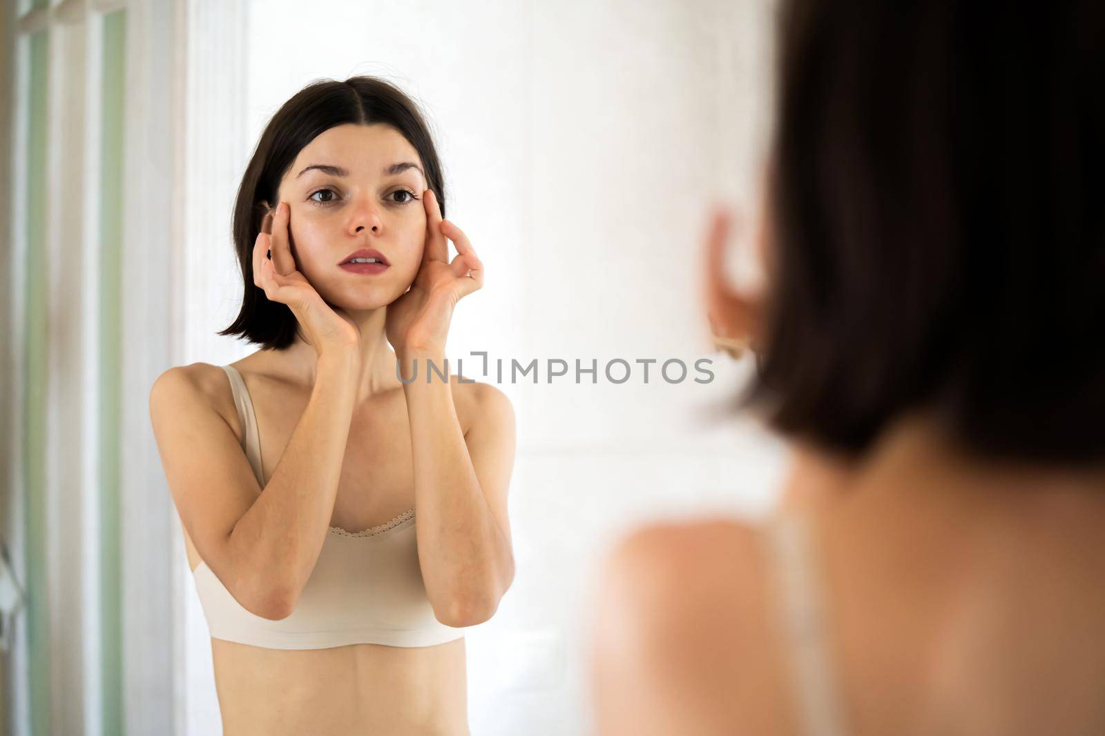 A young girl touches her delicate, clean, moisturized face skin with her hands, a woman takes care of the health and beauty of her body in the bathroom.