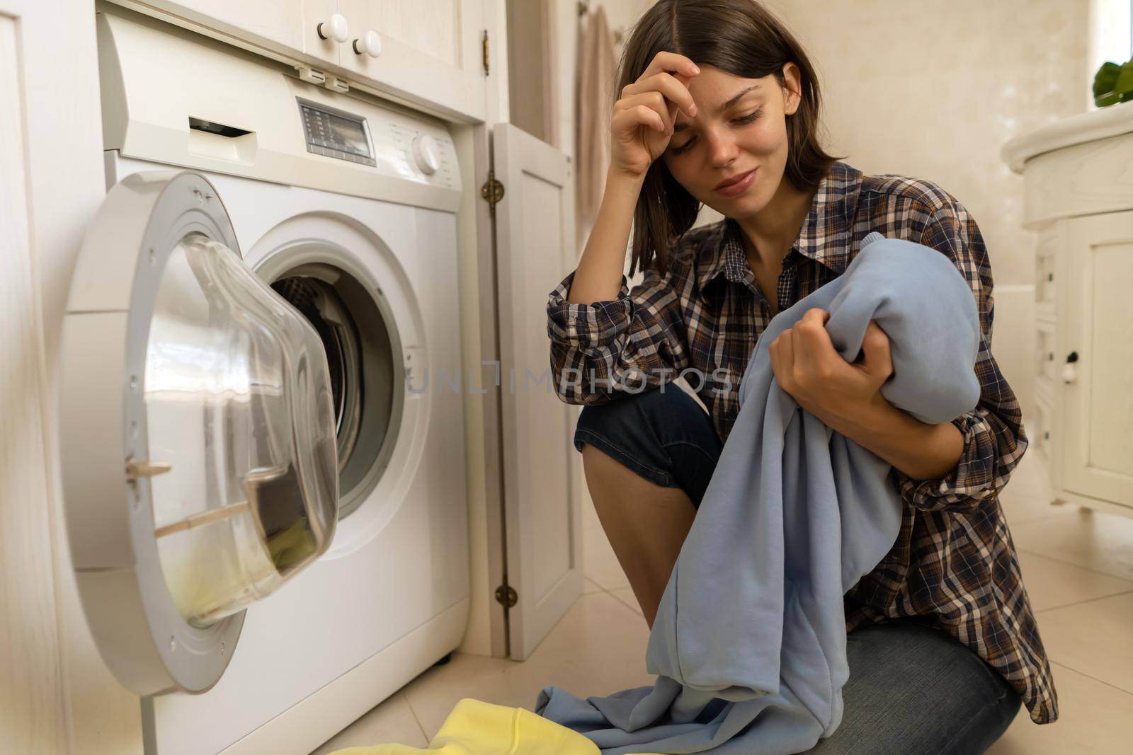 A young girl puts things in the washing machine, a tired woman sits on the floor of a cozy house and looks at the stain on her jacket, which needs to be removed with detergent.