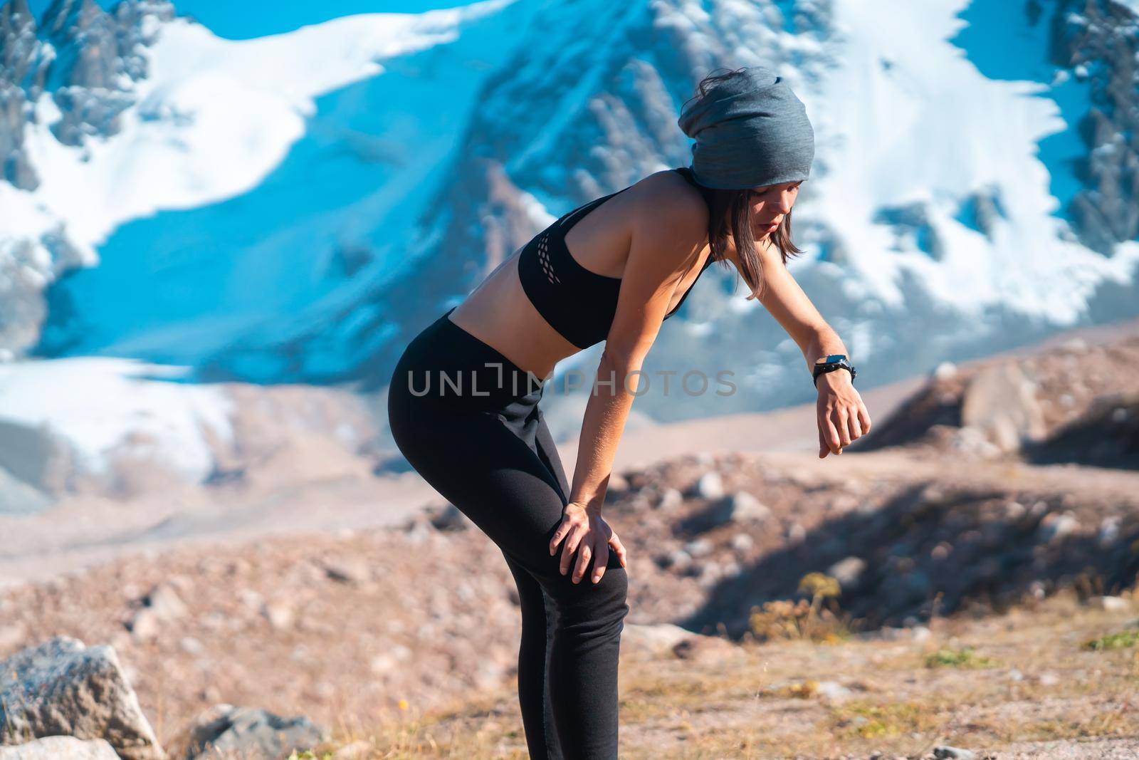 A young athletic girl looks at a smartwatch on her hand, checks her pulse, takes a break after jogging on trails in the snowy mountains. Woman runs outdoors in a mountainous beautiful area.