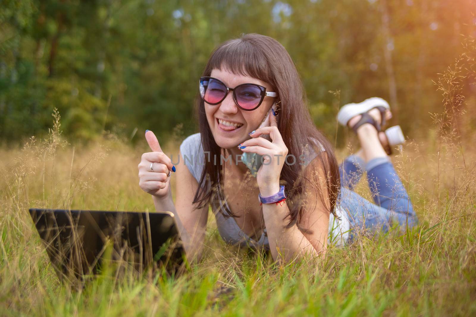 young woman lies on the grass with a laptop and speaks emotionally on the phone