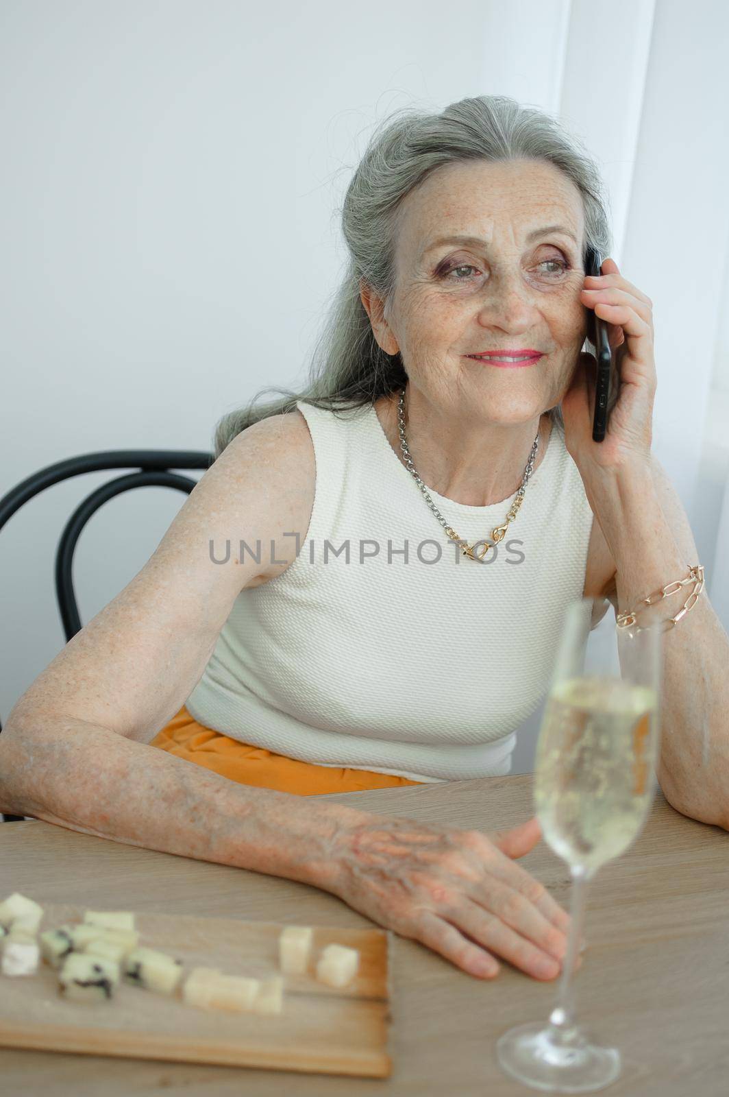 Beautiful old grandmother with grey hair and face with wrinkles is using smartphone, talking with someone and sitting at the table at home on window background, mother's day, happy retirement by balinska_lv