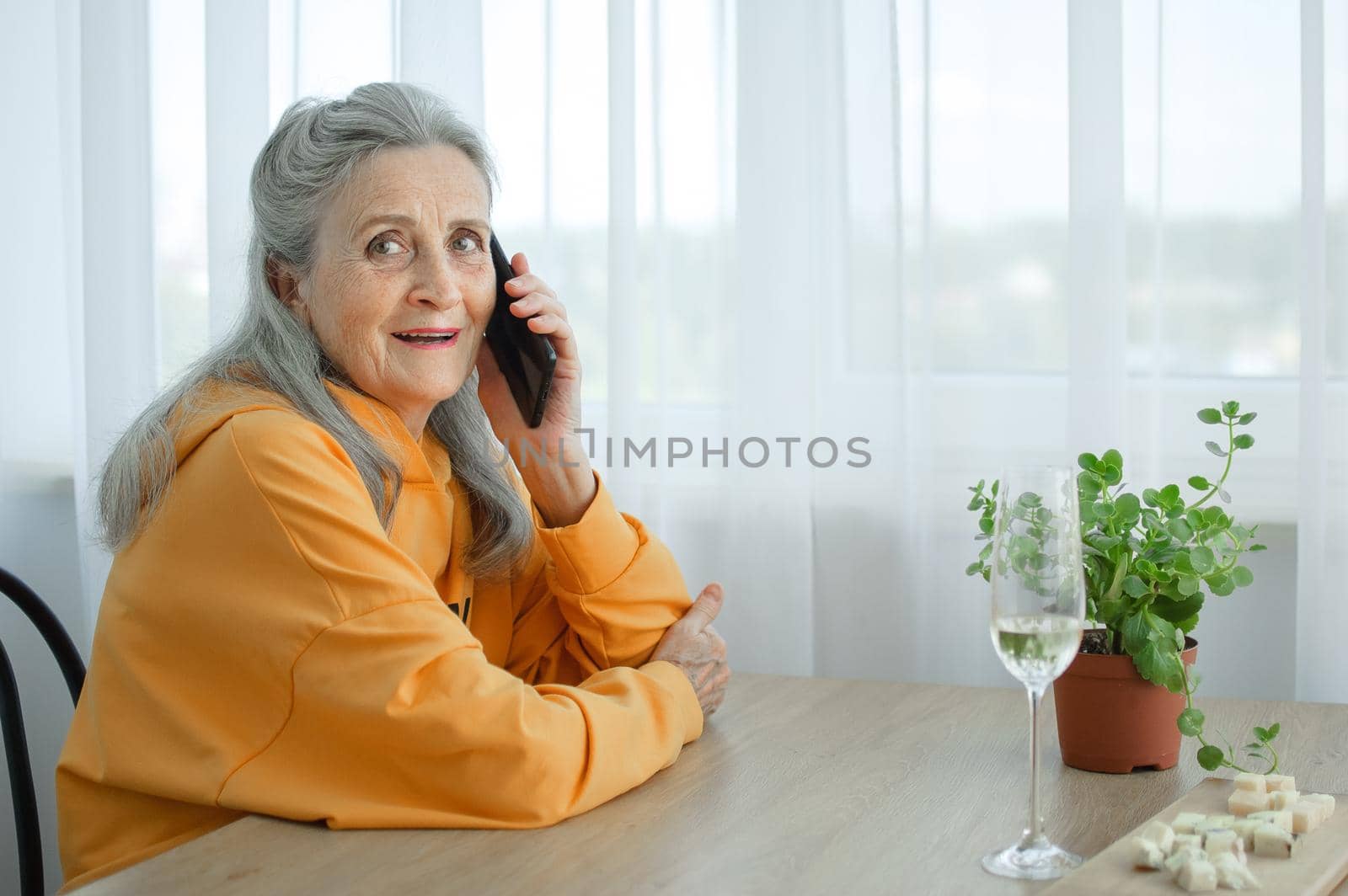 Beautiful old grandmother with grey hair and face with wrinkles is using smartphone, talking with someone and sitting at the table at home on window background, mother's day, happy retirement.