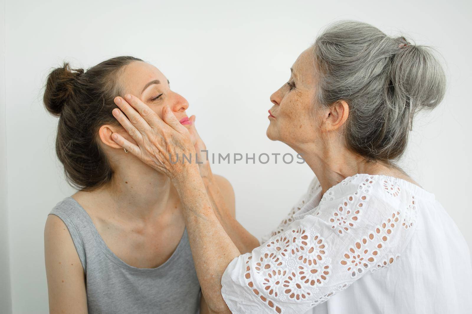 Happy senior mother is hugging her adult daughter, the women are laughing together, sincere family of different age generations having fun on white background by balinska_lv