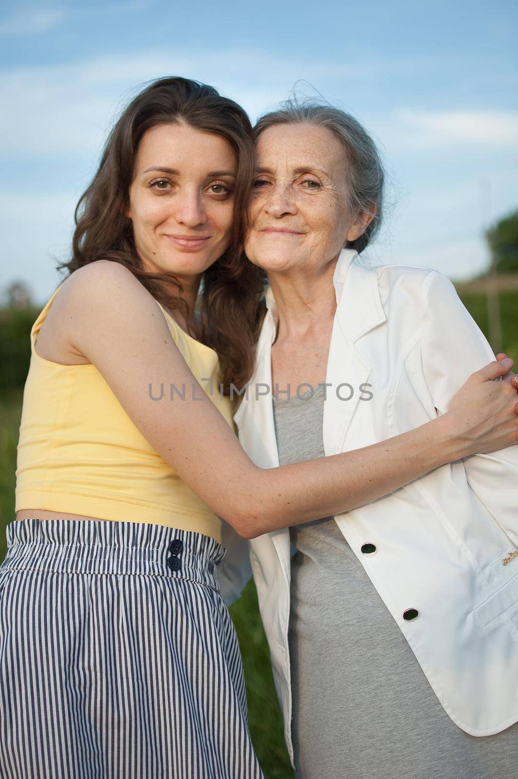 Senior mother with gray hair with her adult daughter looking at the camera in the garden and hugging each other during sunny day outdoors, mothers day concept