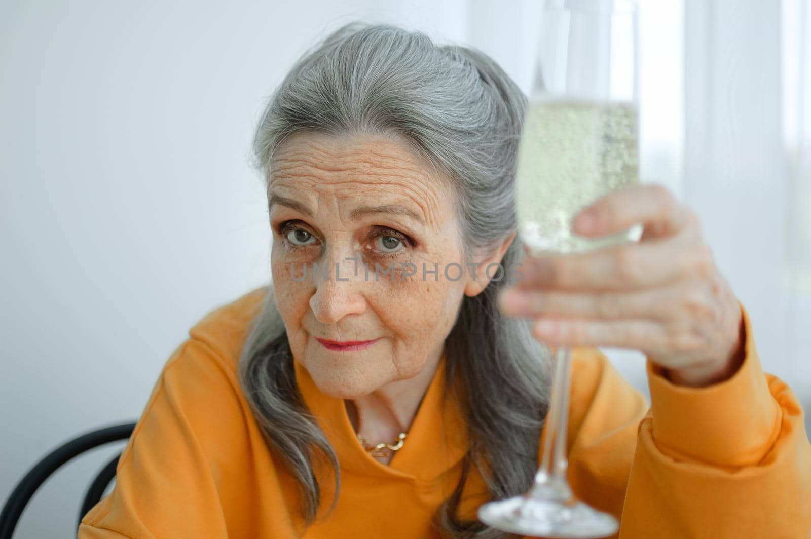 Beautiful old grandmother with grey hair and face with wrinkles sitting at the table at home on window background with glass of champagne, mother's day, happy retirement by balinska_lv