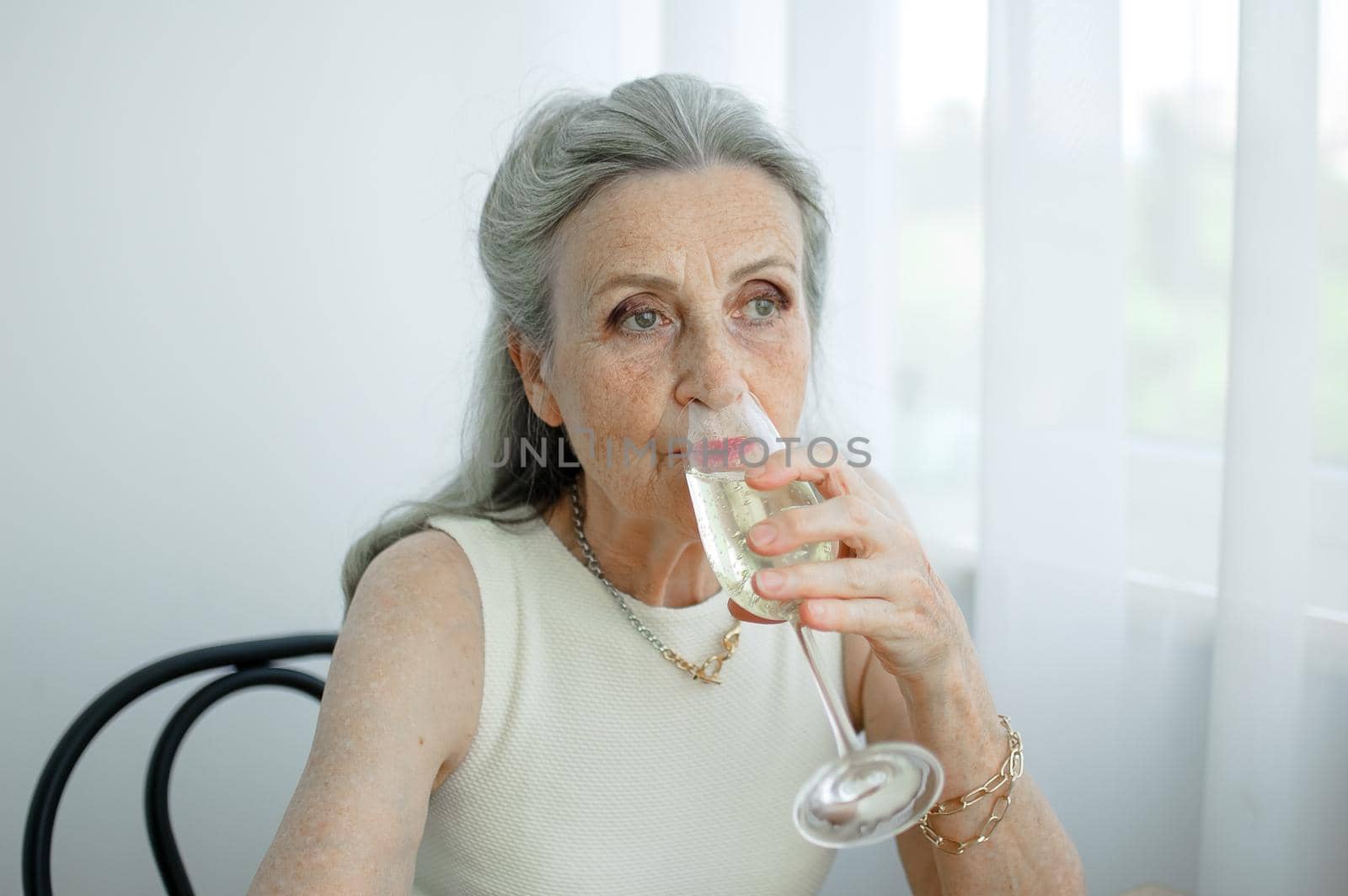 Beautiful old grandmother with grey hair and face with wrinkles sitting at the table at home on window background with glass of champagne, mother's day, happy retirement.