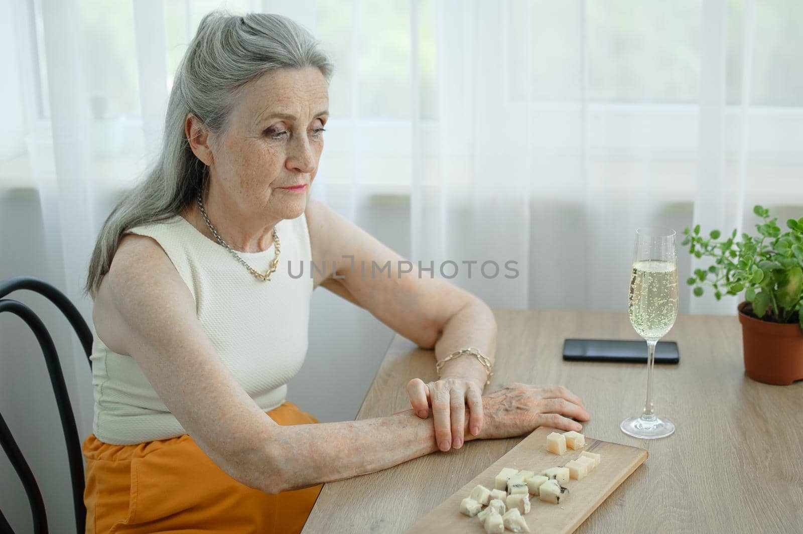 Beautiful old grandmother with grey hair and face with wrinkles sitting at the table at home on window background with glass of champagne, mother's day, happy retirement.